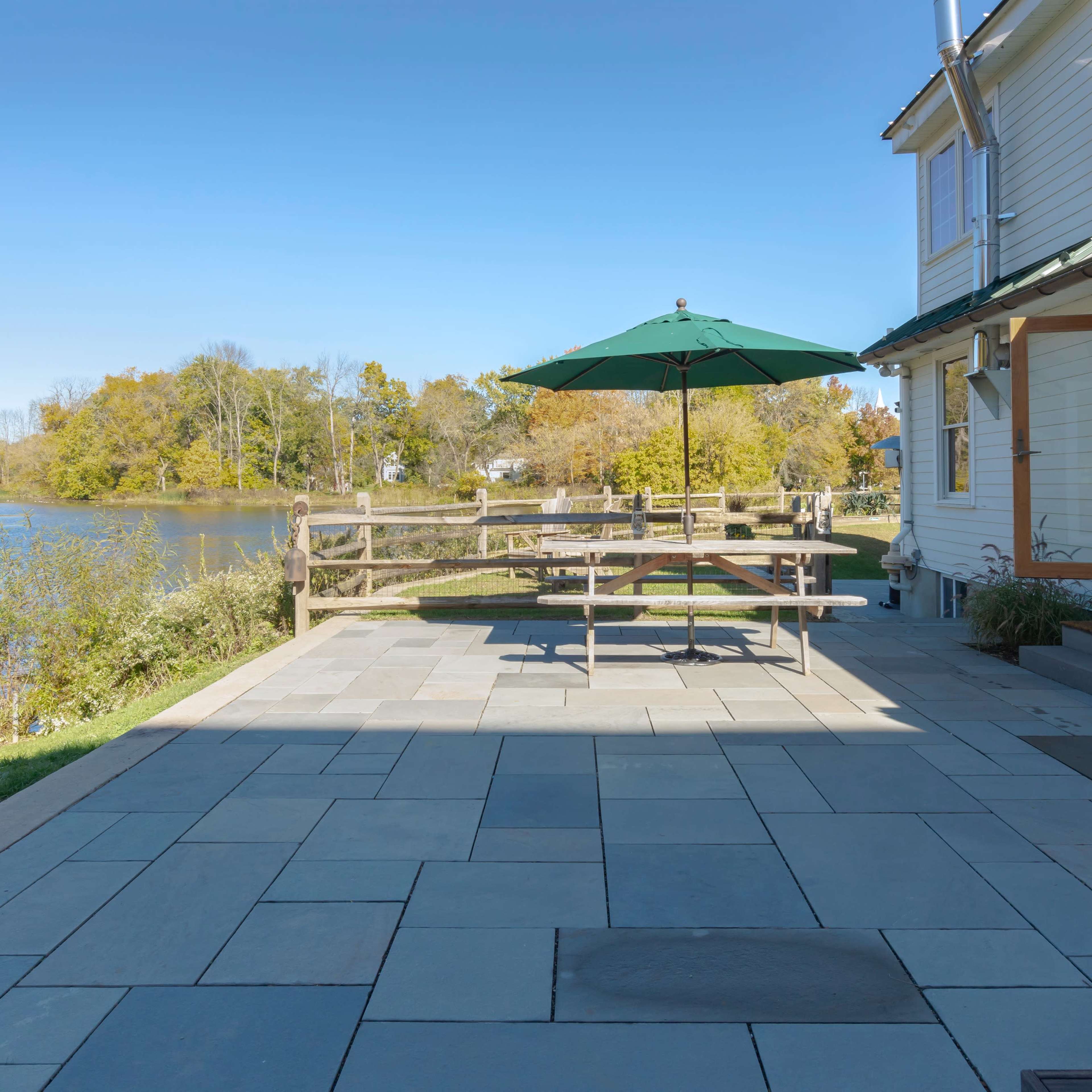 A patio with a wooden table and a green umbrella overlooks a calm body of water surrounded by trees.