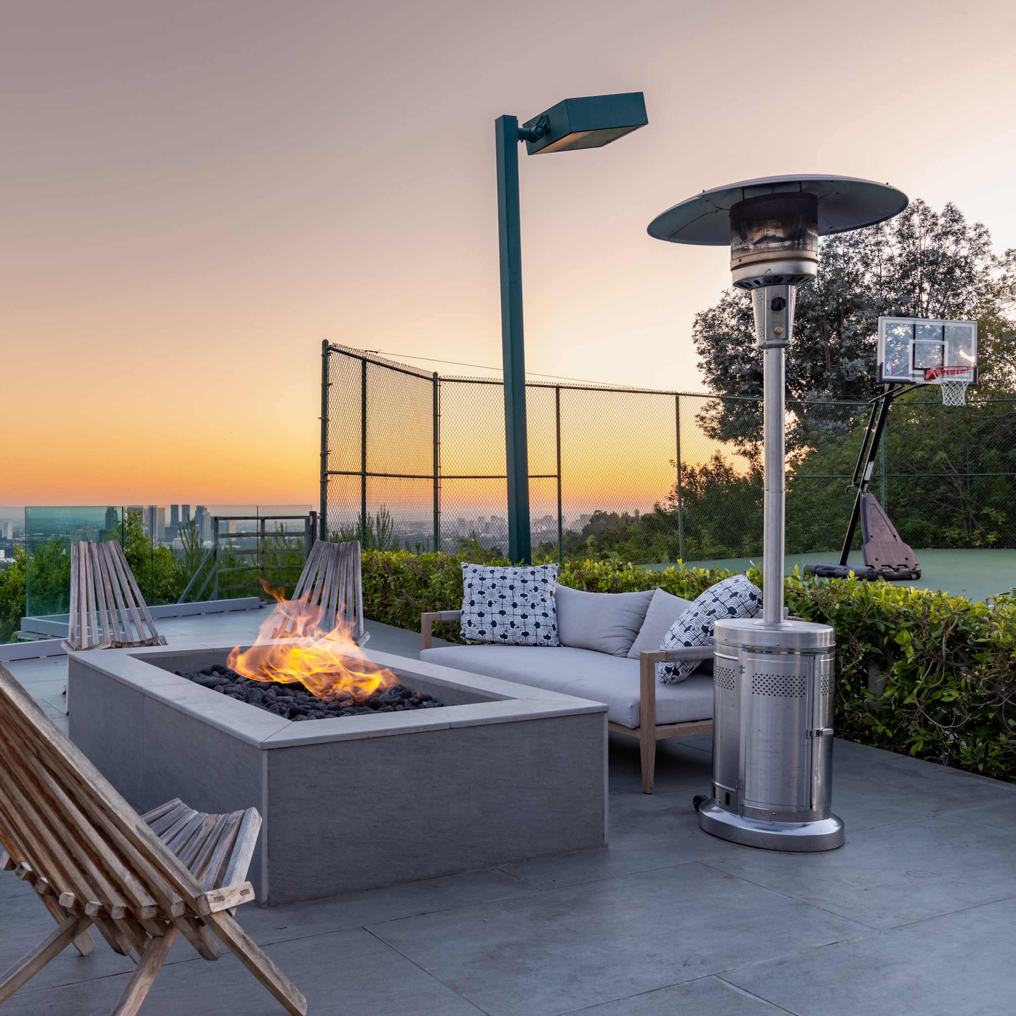 A modern outdoor seating area features a fire pit surrounded by chairs, with a view of the sunset and a basketball hoop in the background.