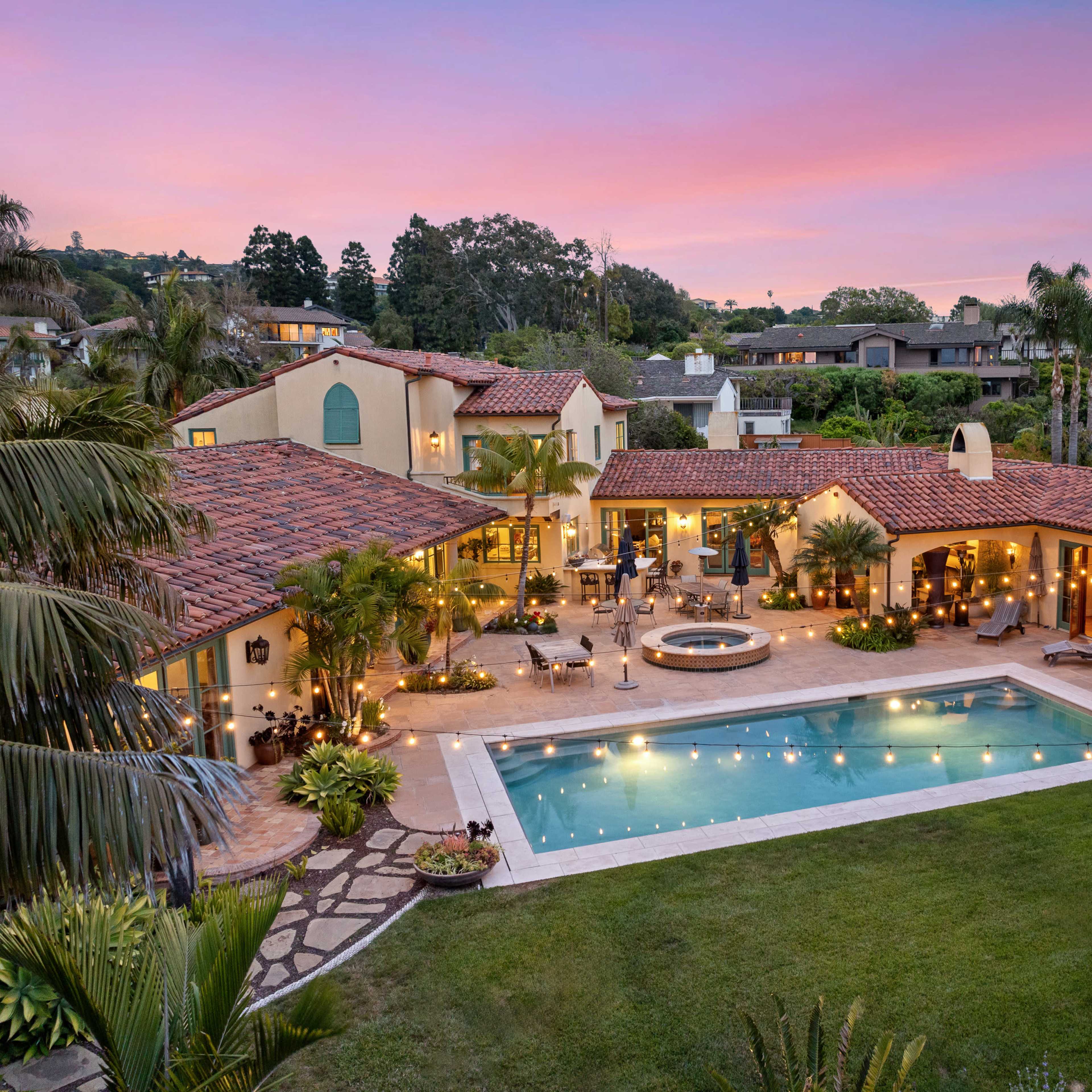 A large, well-landscaped backyard features a pool, spa, and seating area surrounded by palm trees and illuminated by string lights, with a spacious home in the background.