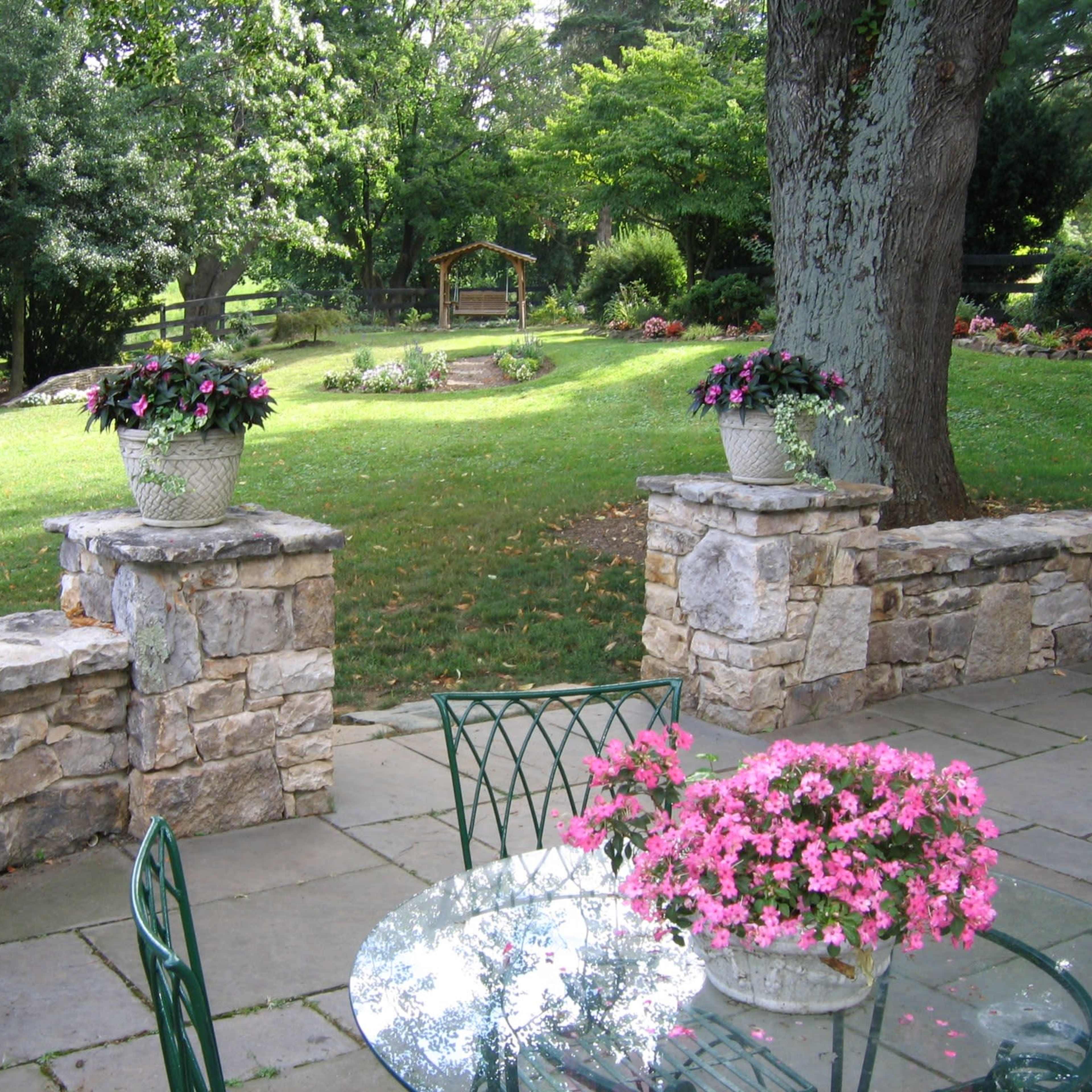 A stone patio with potted flowers and a view of a landscaped garden featuring a wooden swing in the background.