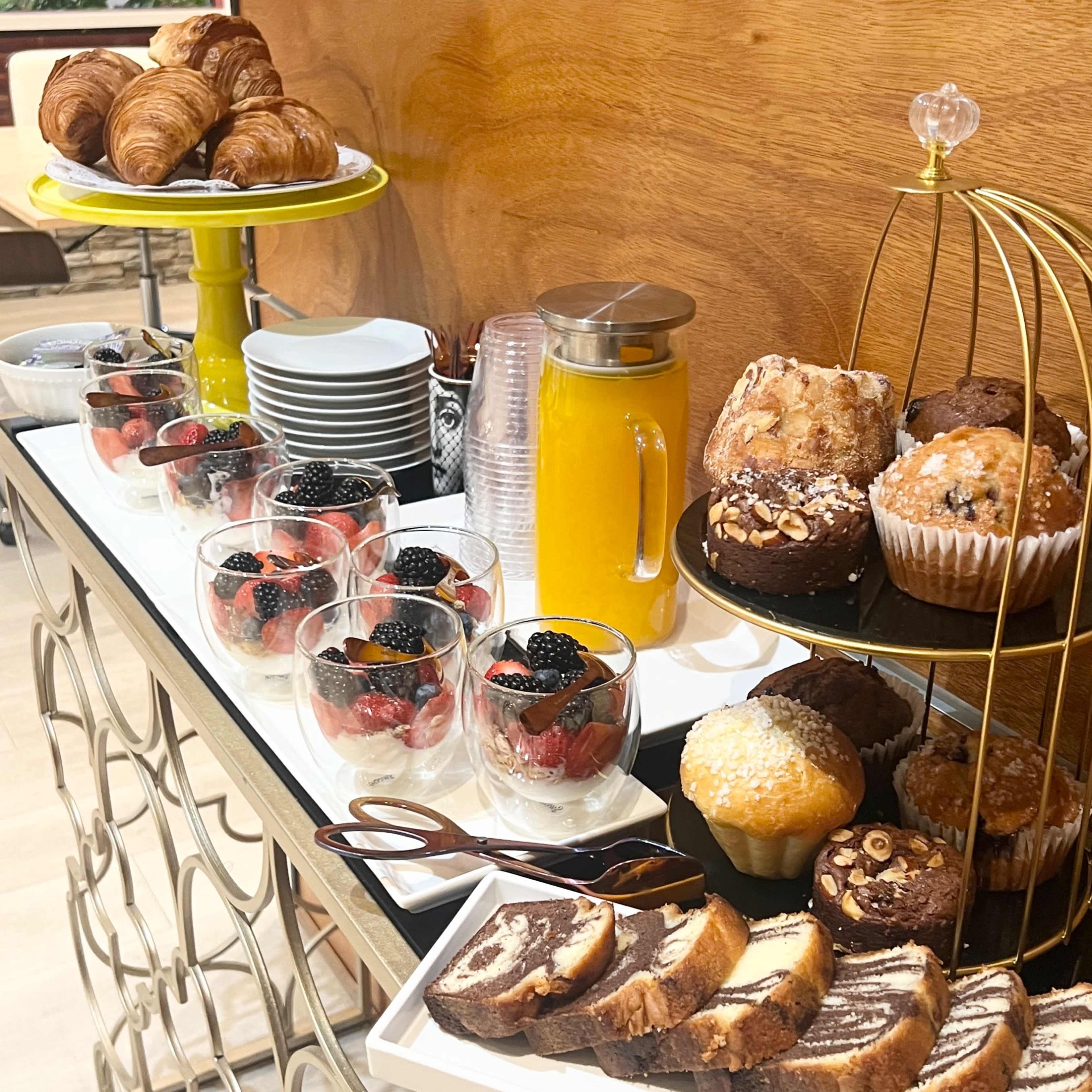 A dessert table features croissants, muffins, chocolate cake, fruit cups with berries, and a pitcher of orange juice.