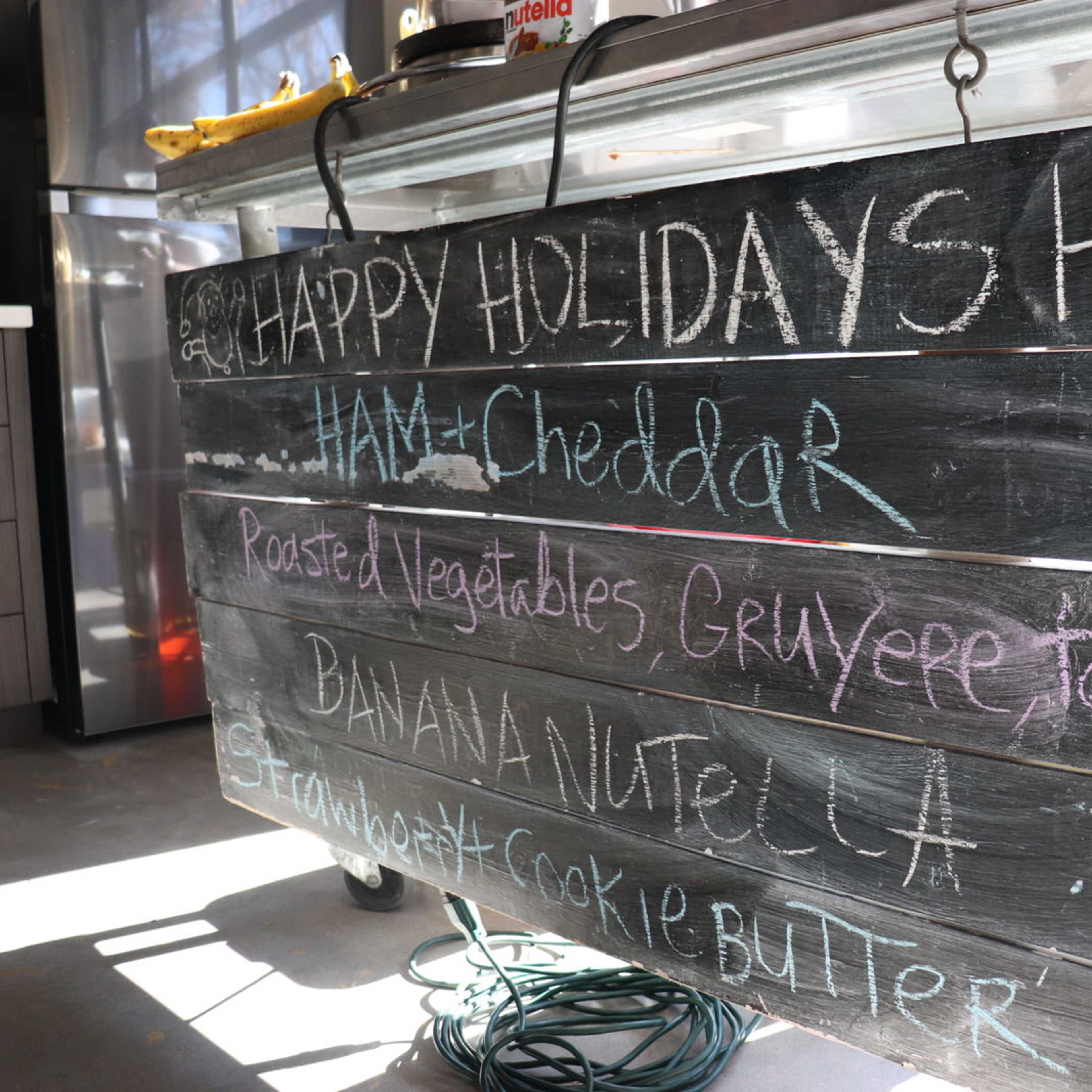 A chalkboard menu displays various food items, including ham and cheddar, roasted vegetables, banana and Nutella, and strawberry cookie butter, along with a holiday greeting.