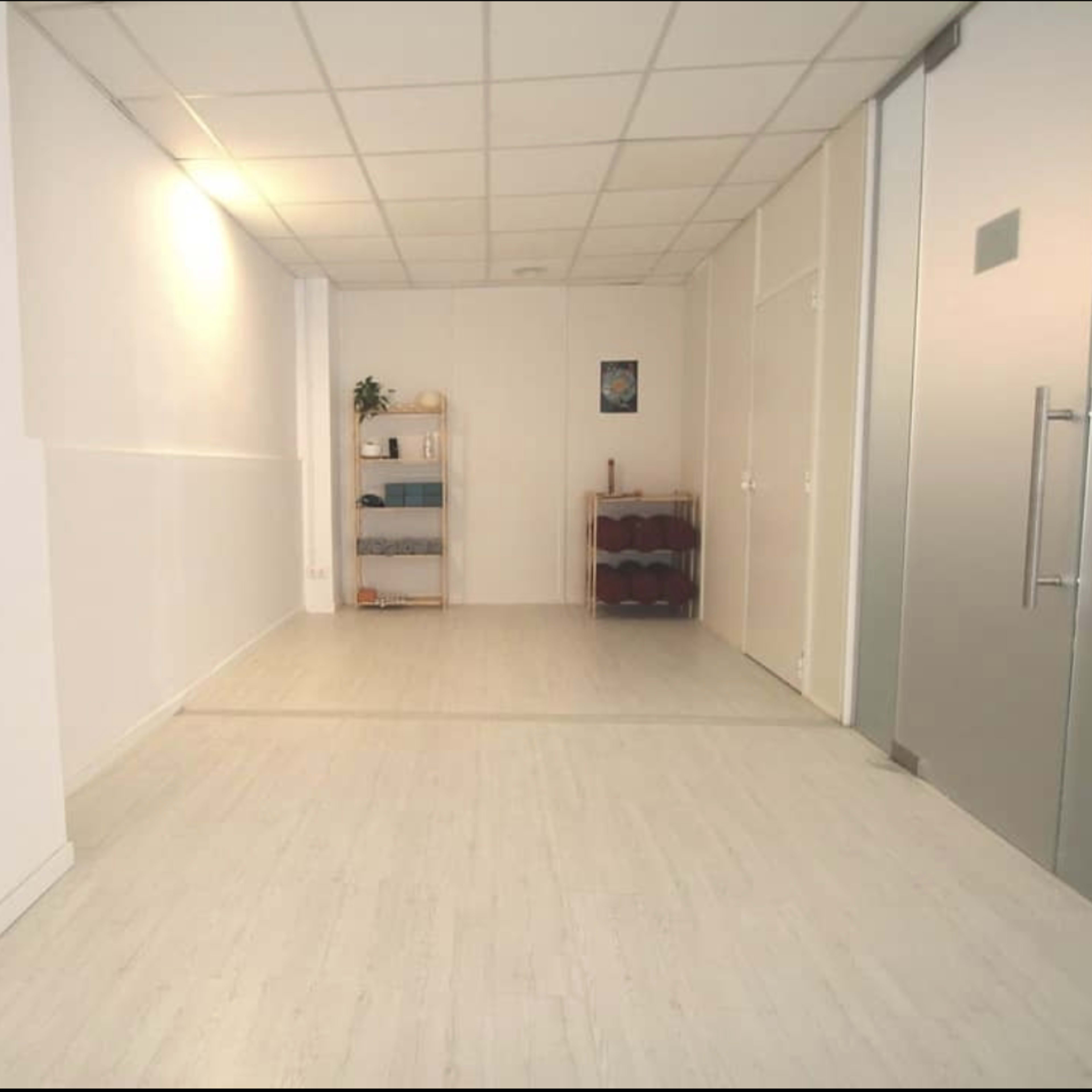 An empty, well-lit room with white walls and a light-colored floor, featuring a shelf against one wall and a closed glass door on the right.