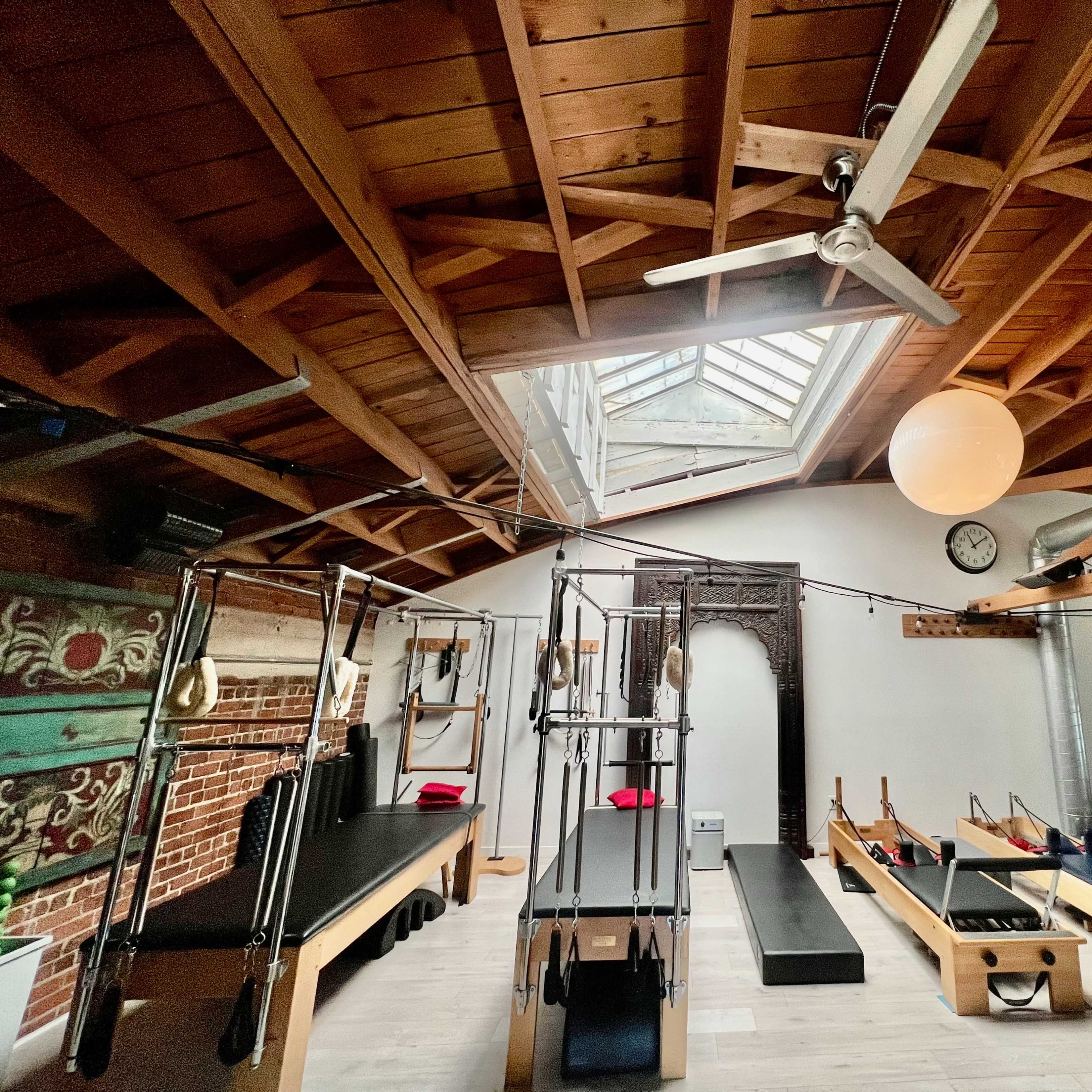 A spacious Pilates studio with reformer machines lined along the walls, wooden beams overhead, and a skylight illuminating the space.