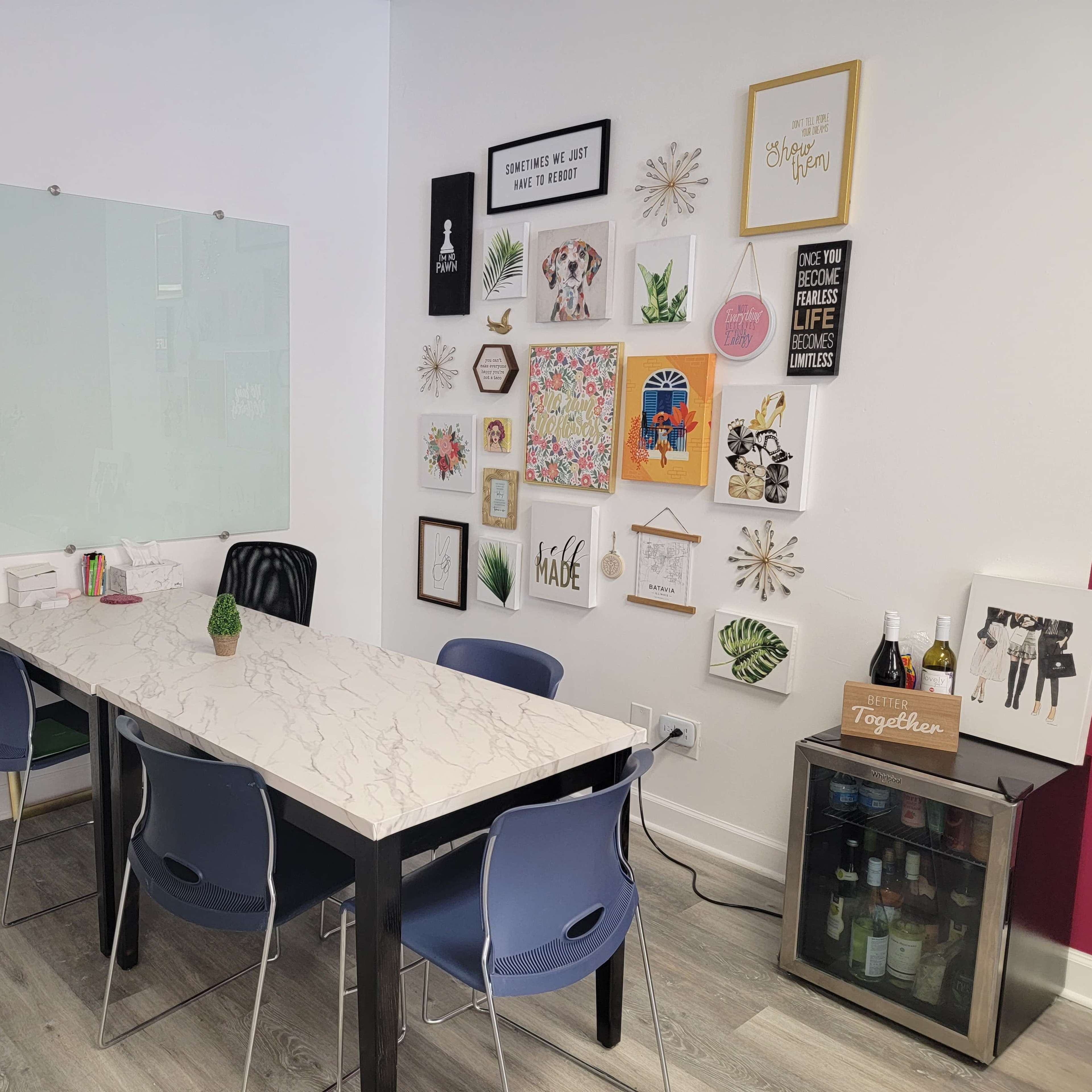 A modern meeting room with a marble table, blue chairs, a glass whiteboard, and a wall decorated with various framed artworks and a small beverage refrigerator.