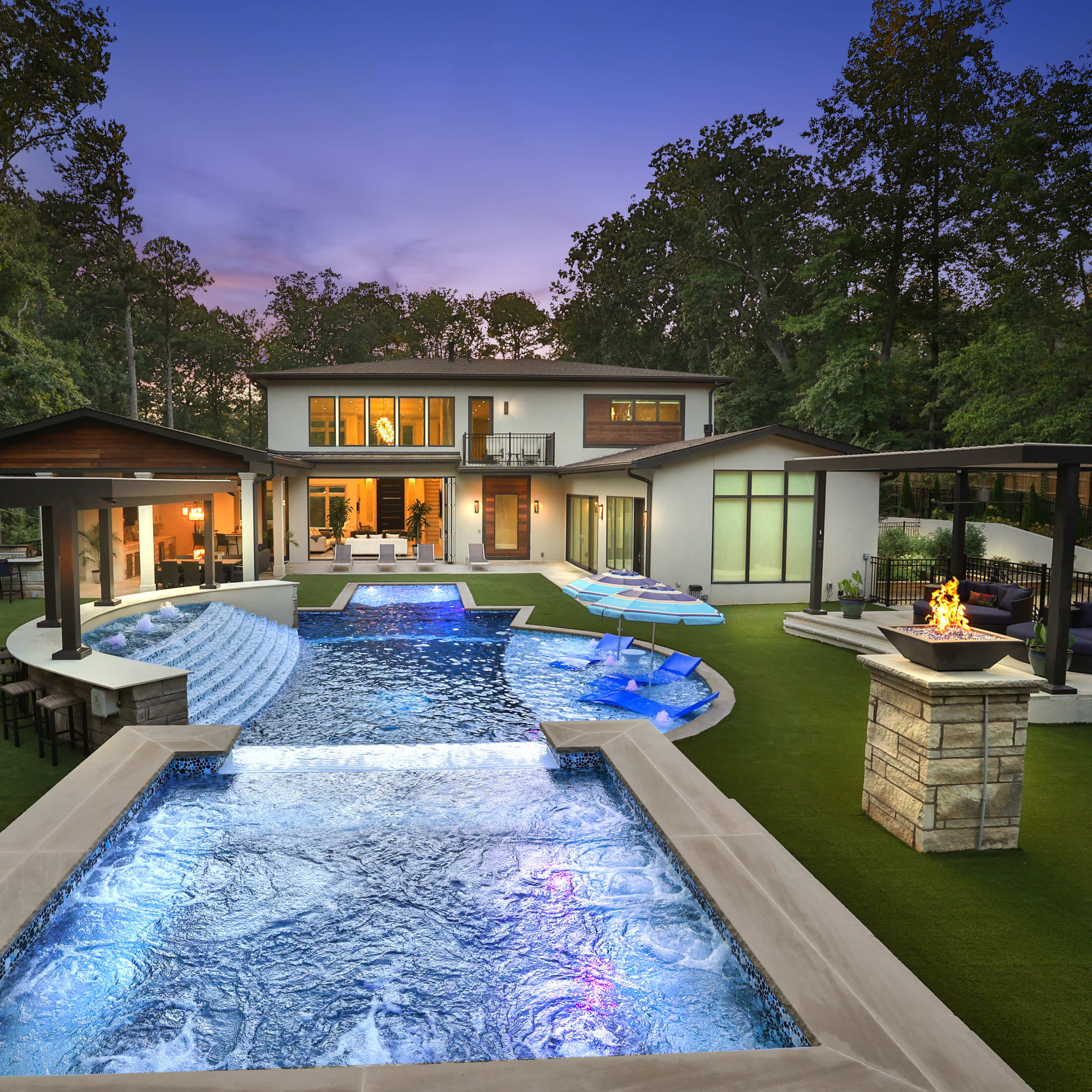 A modern house with a large swimming pool and outdoor seating area, surrounded by trees at dusk.
