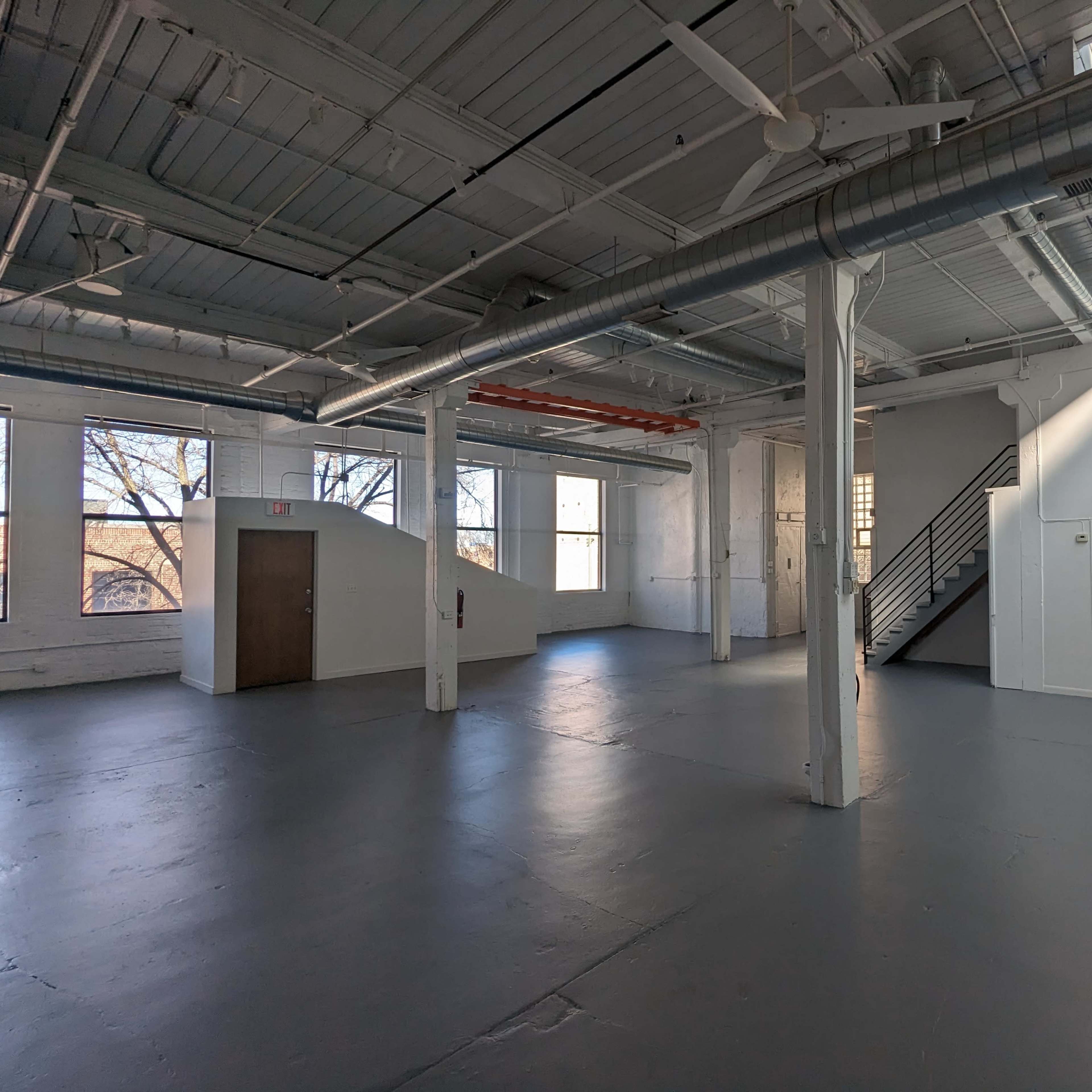 A spacious, well-lit industrial-style room with large windows and a staircase in the corner.