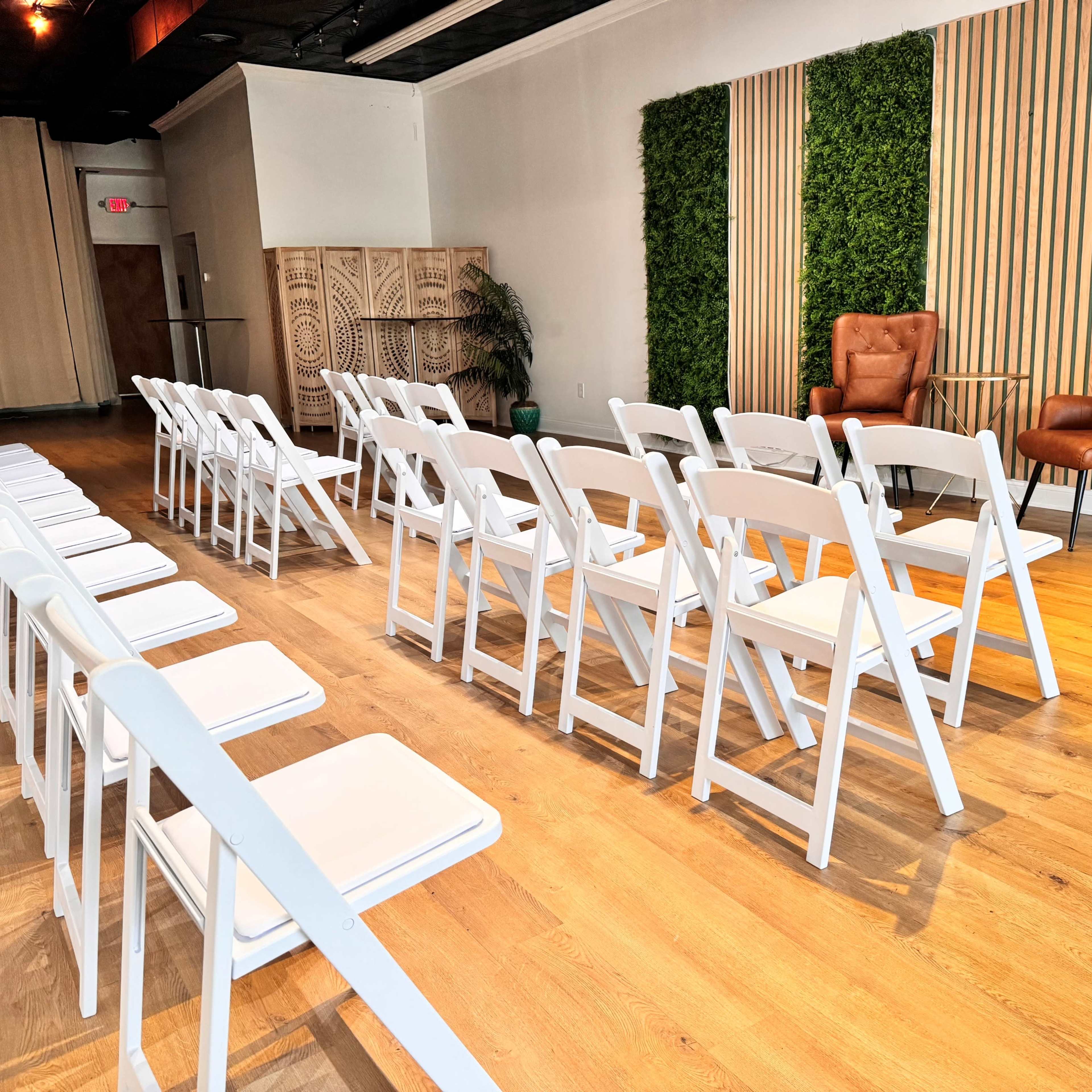 A row of white folding chairs arranged in a spacious room with a green wall and two brown armchairs set against a wooden backdrop.
