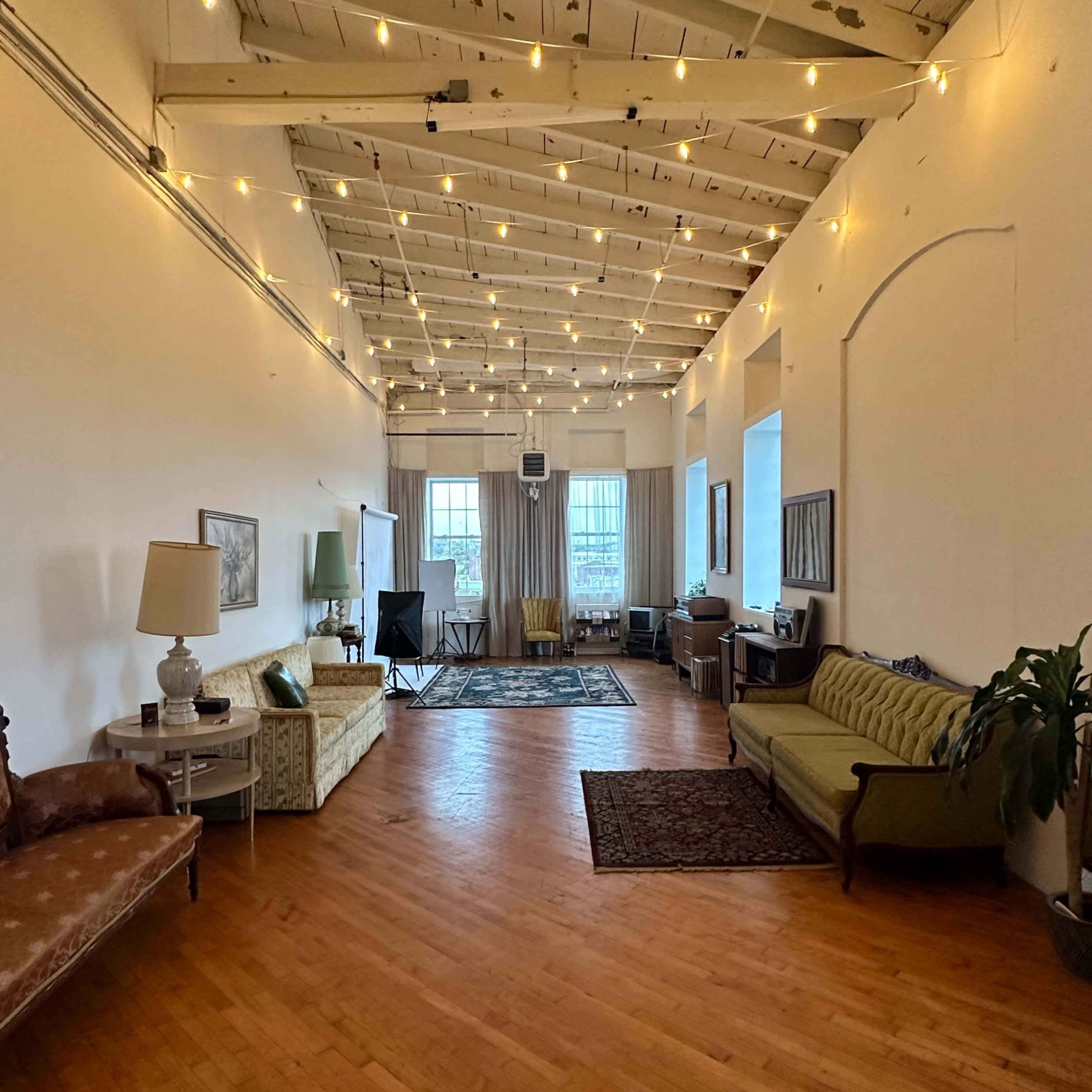A spacious, well-lit room with wooden flooring, string lights hanging from the ceiling, and a mix of vintage furniture along the walls.