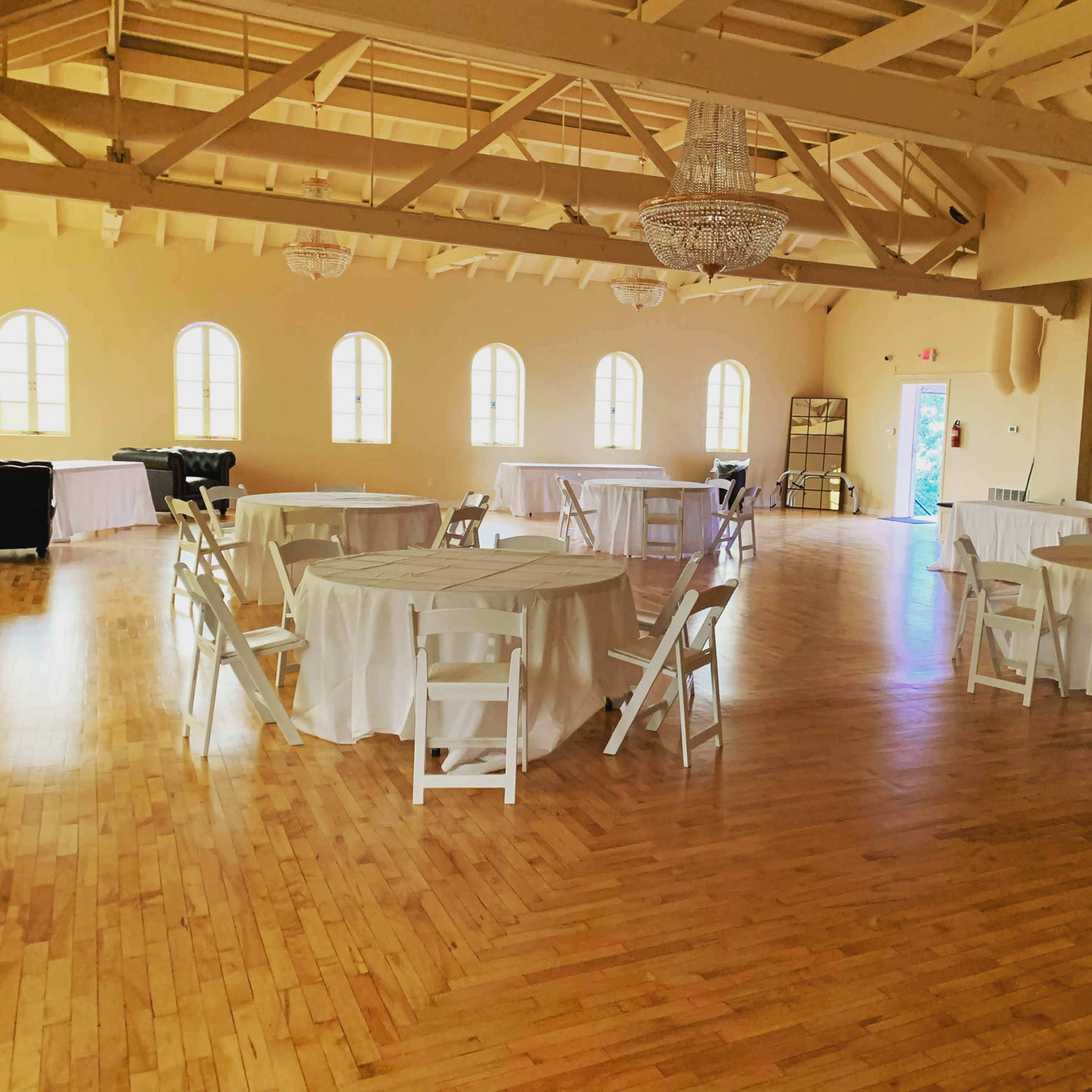 A spacious event hall with wooden flooring, round tables covered in white cloth, and large windows letting in natural light.