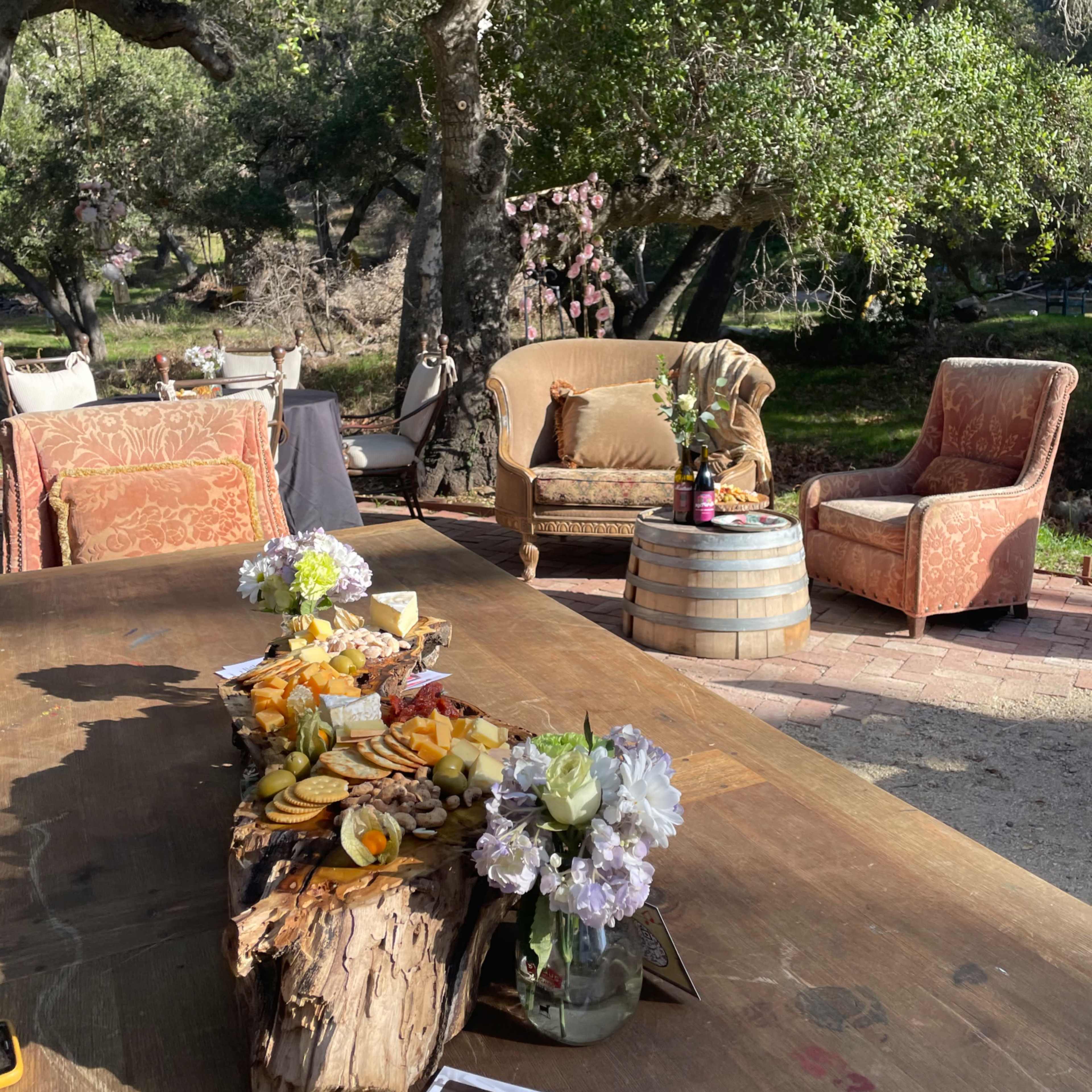 A long wooden table is set outdoors with a fruit and flower arrangement, while two armchairs sit nearby under oak trees.