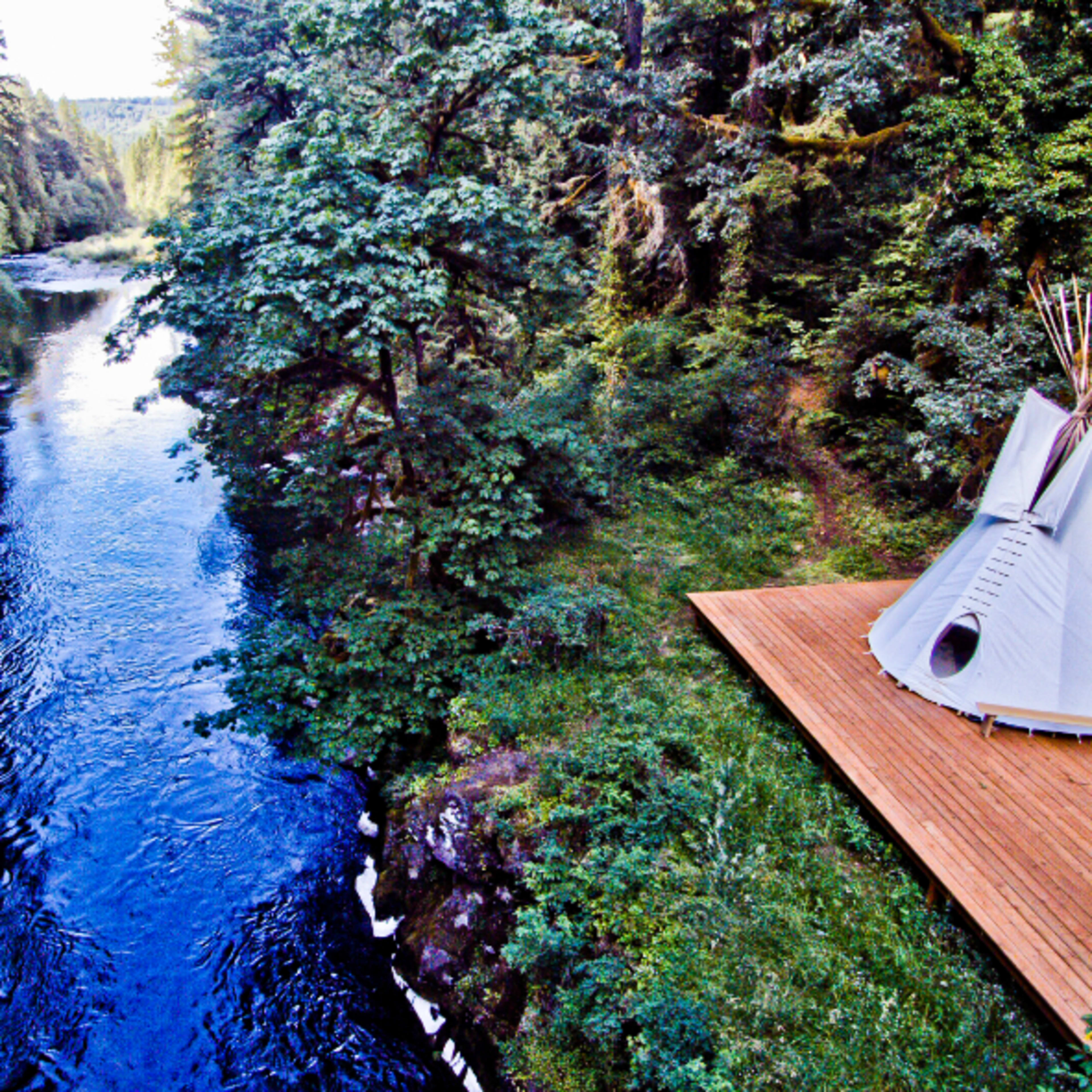 A wooden platform with a teepee overlooks a river surrounded by dense trees.
