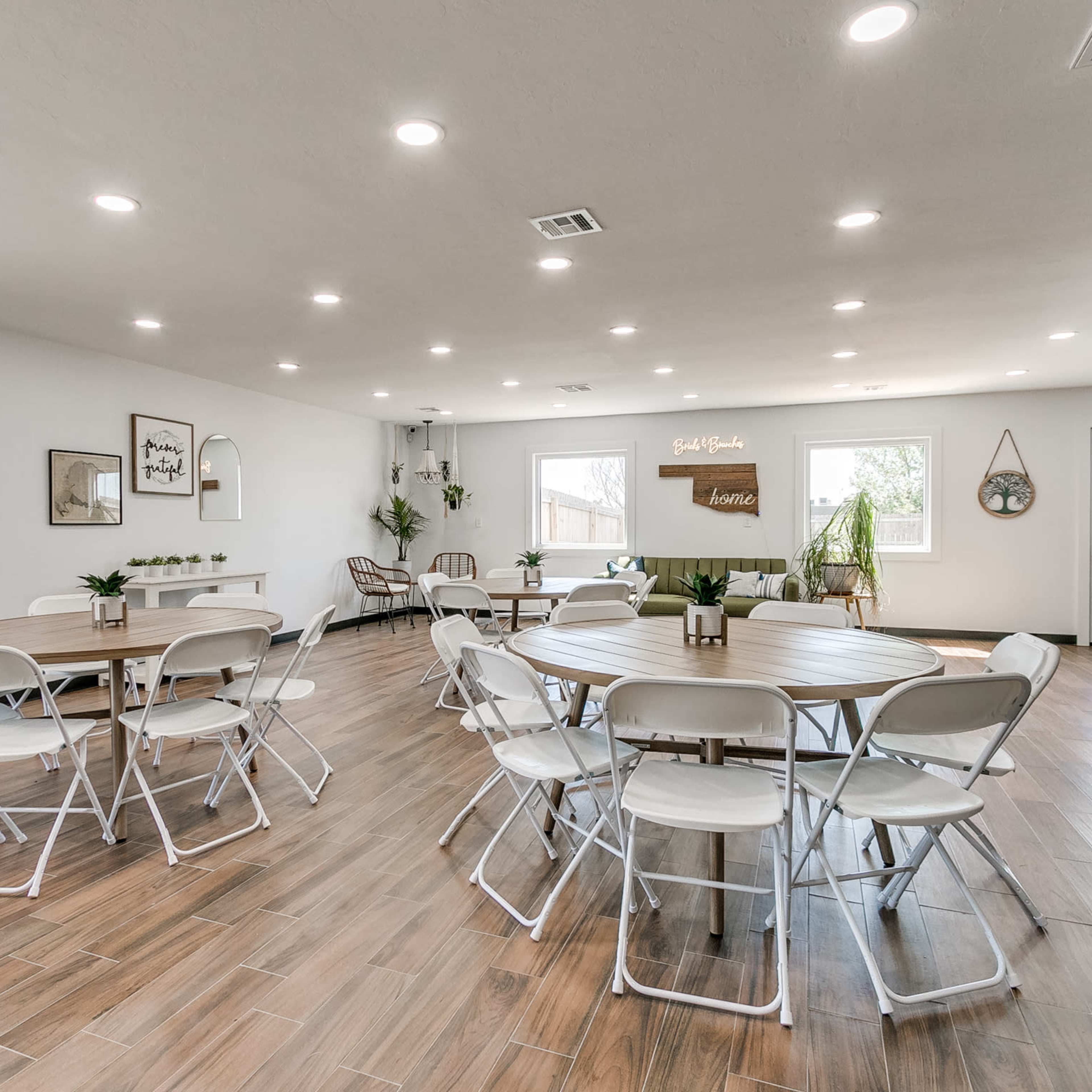 A spacious, well-lit room with multiple round tables and chairs arranged neatly, along with potted plants and a door leading outside.