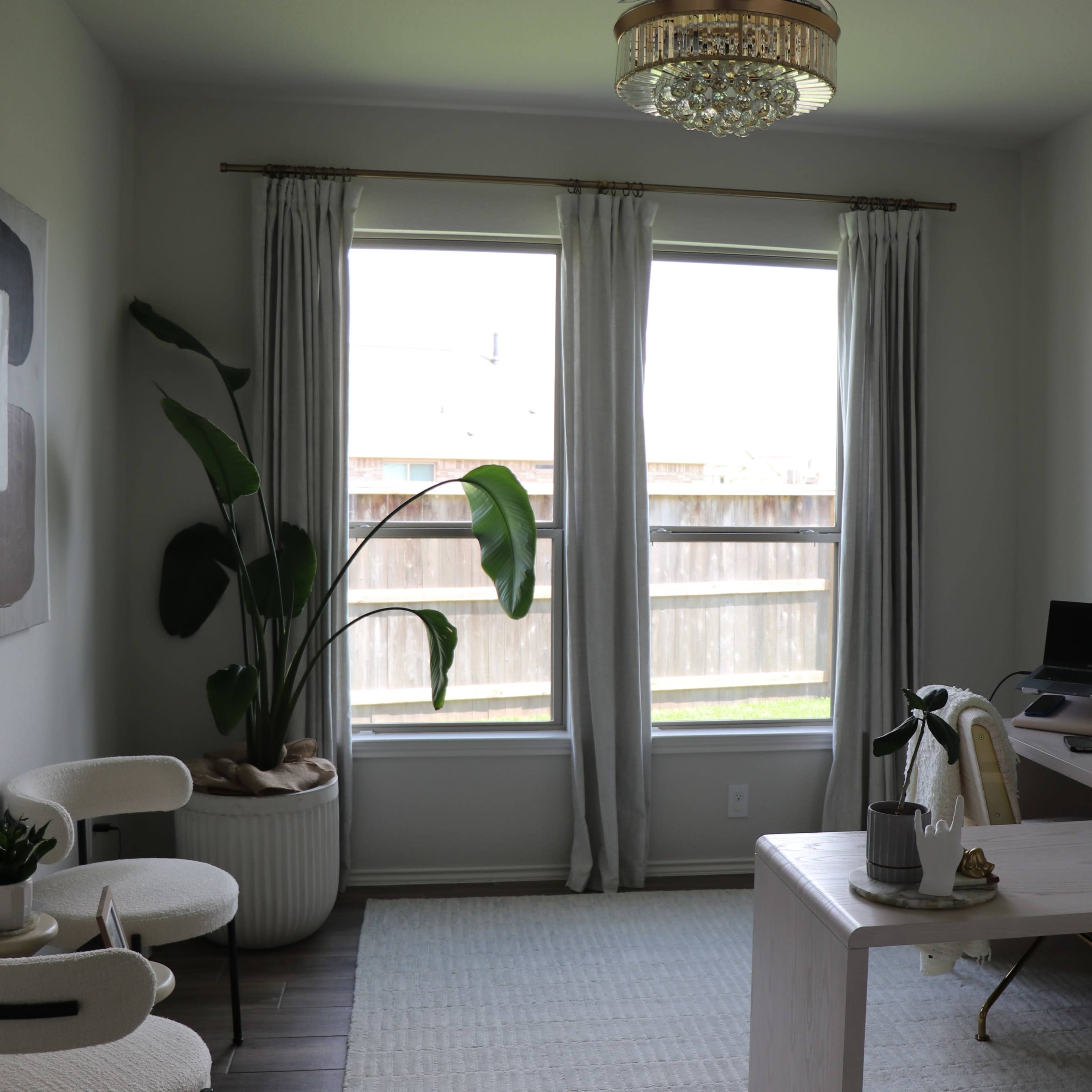 A home office features a desk with two computers, a large plant, and two chairs, illuminated by natural light from two windows.