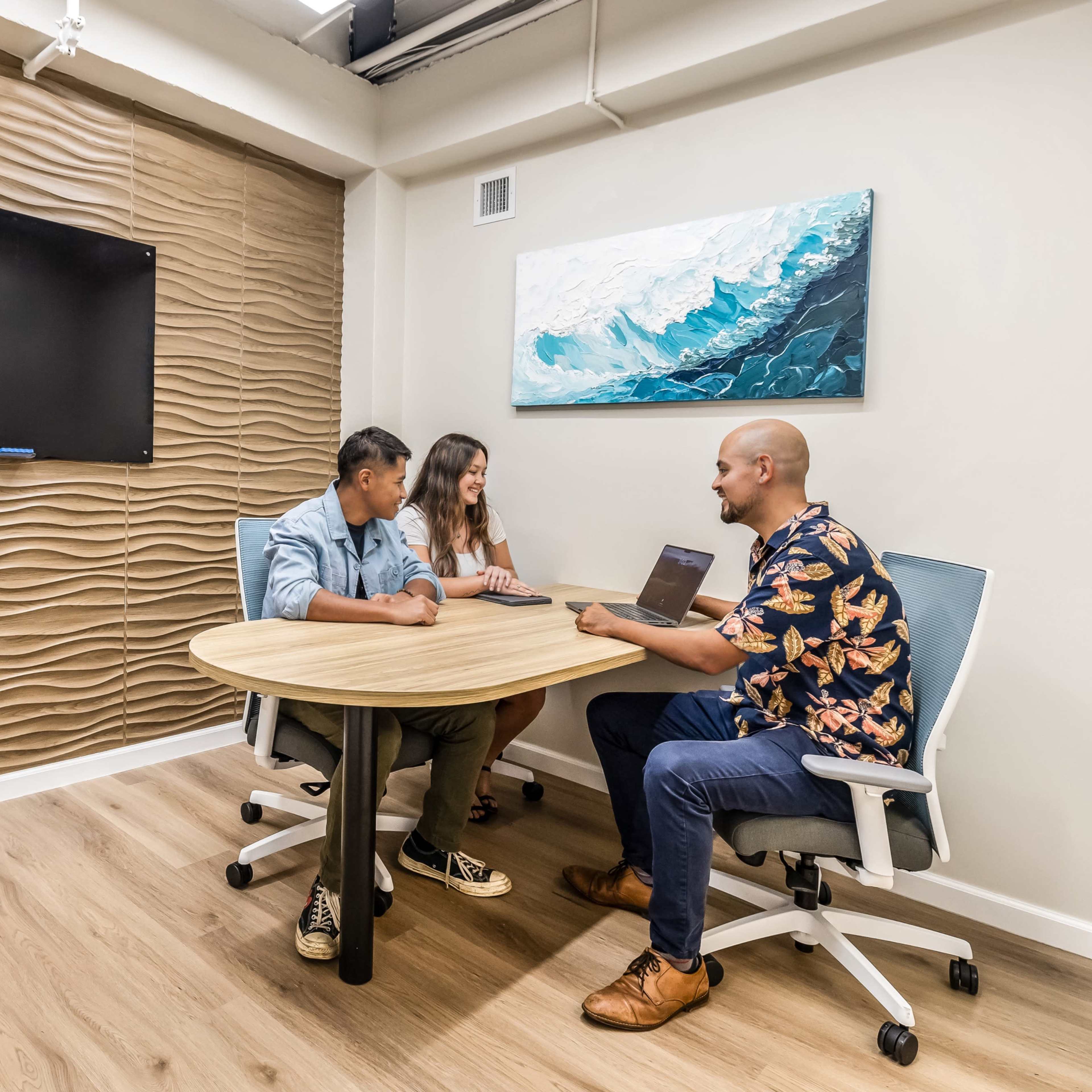 Three people are seated around a round table in a meeting room, with a wall-mounted screen in the background.