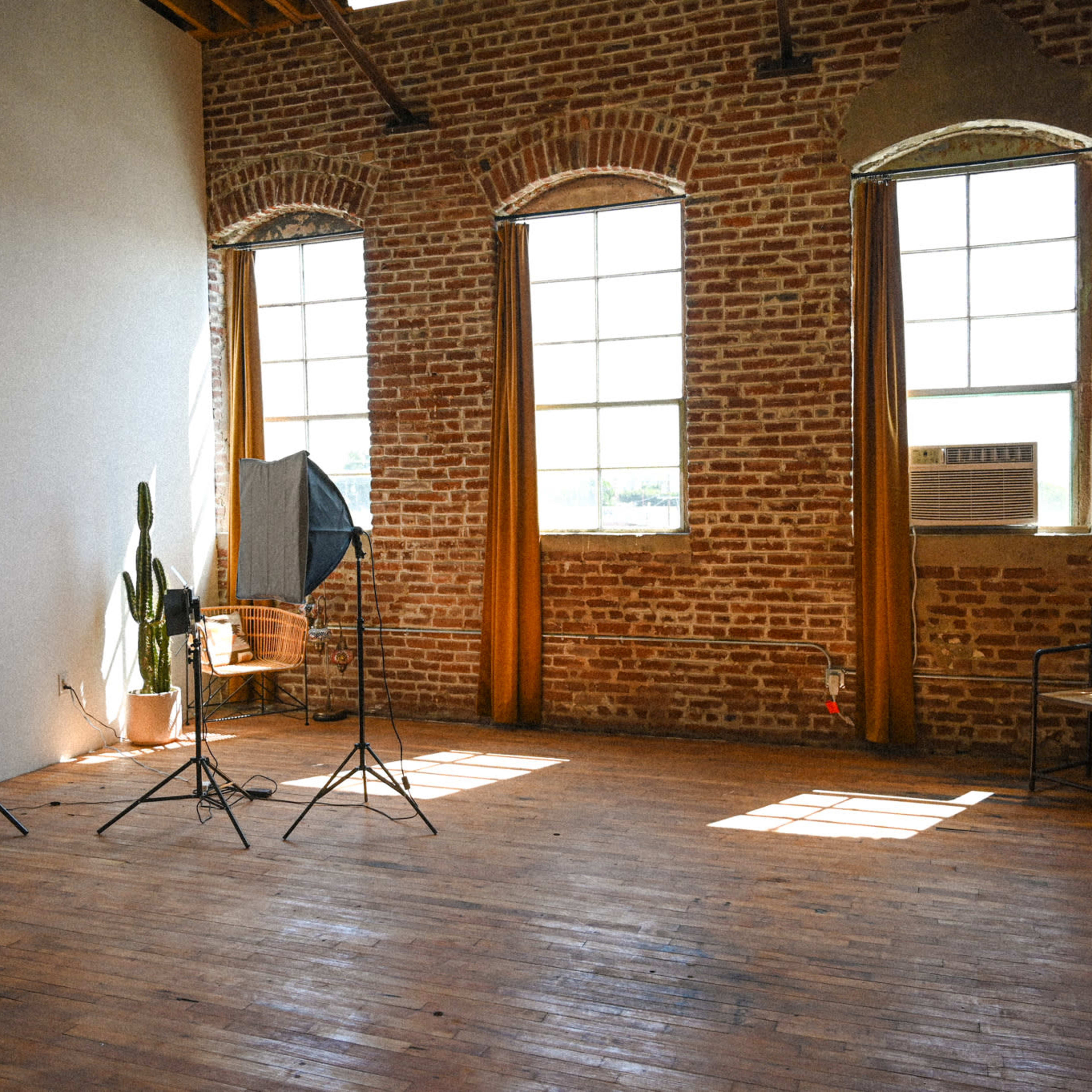 A spacious room with exposed brick walls, large windows, and wooden flooring contains lighting equipment and a potted cactus.