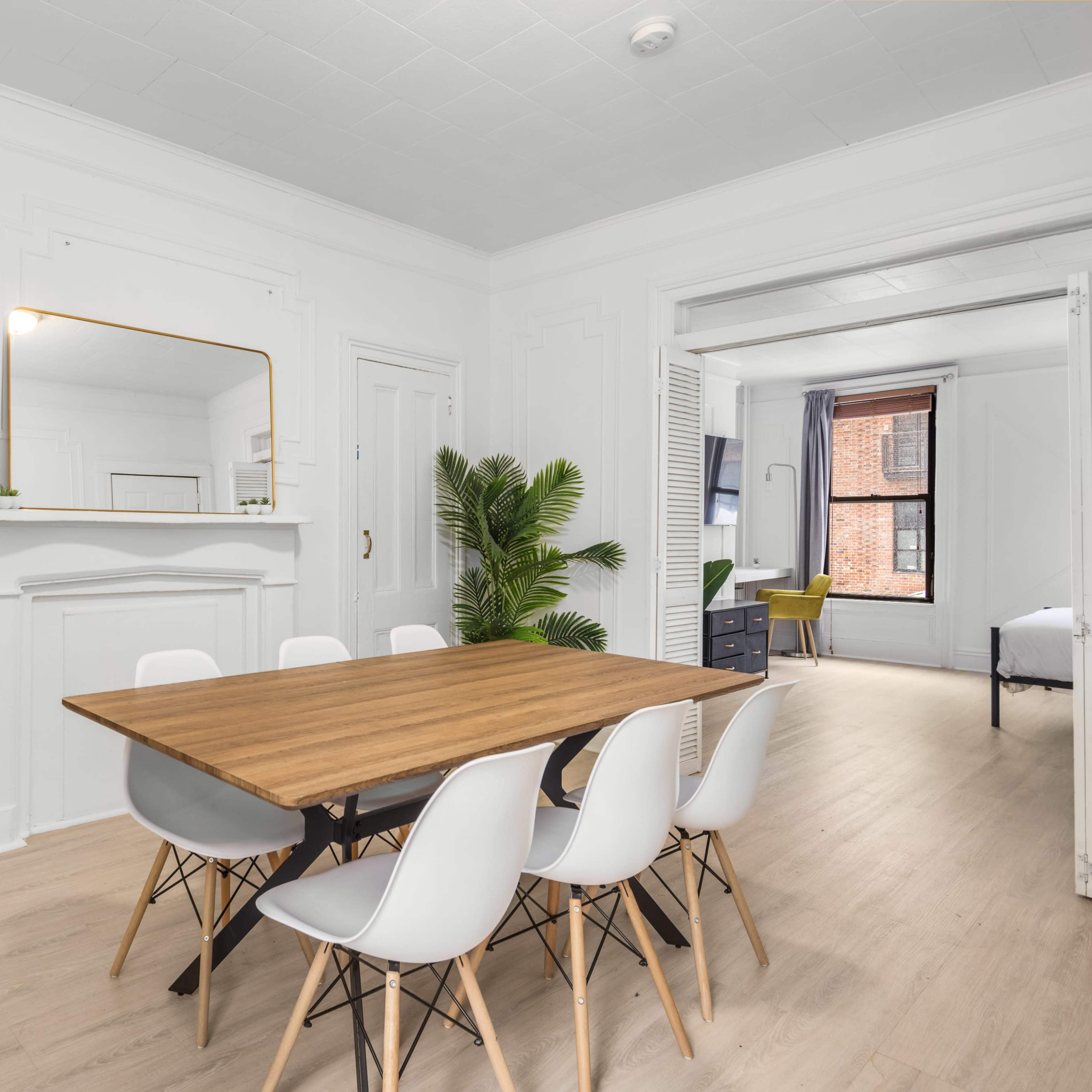 A bright dining area with a wooden table surrounded by white chairs, a golden bar cart in the corner, and a green plant beside a mirror on a wall with decorative moldings.