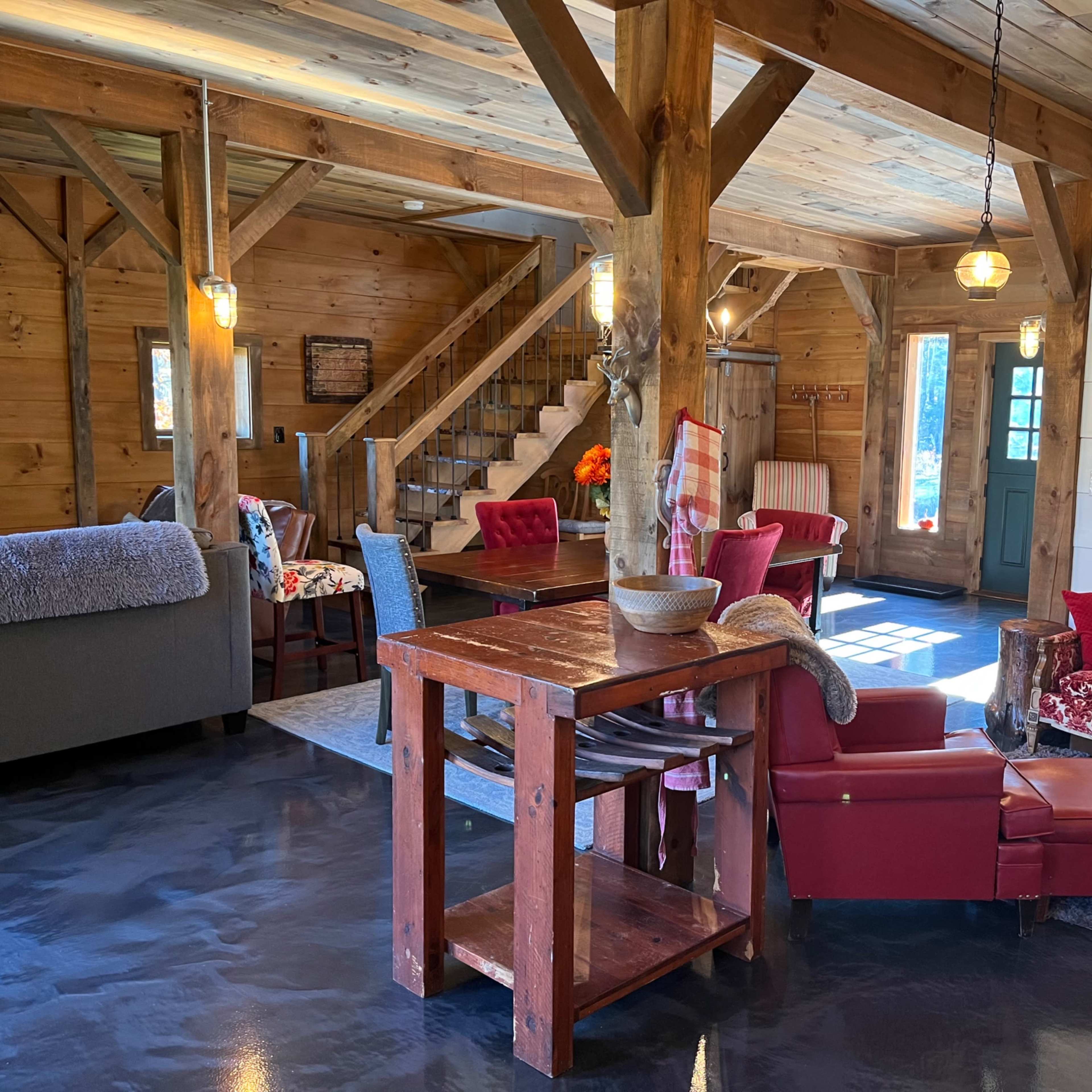 A bright, rustic interior of a wooden cabin featuring a mix of red and cream furniture, a dining area, and a staircase leading to an upper level.