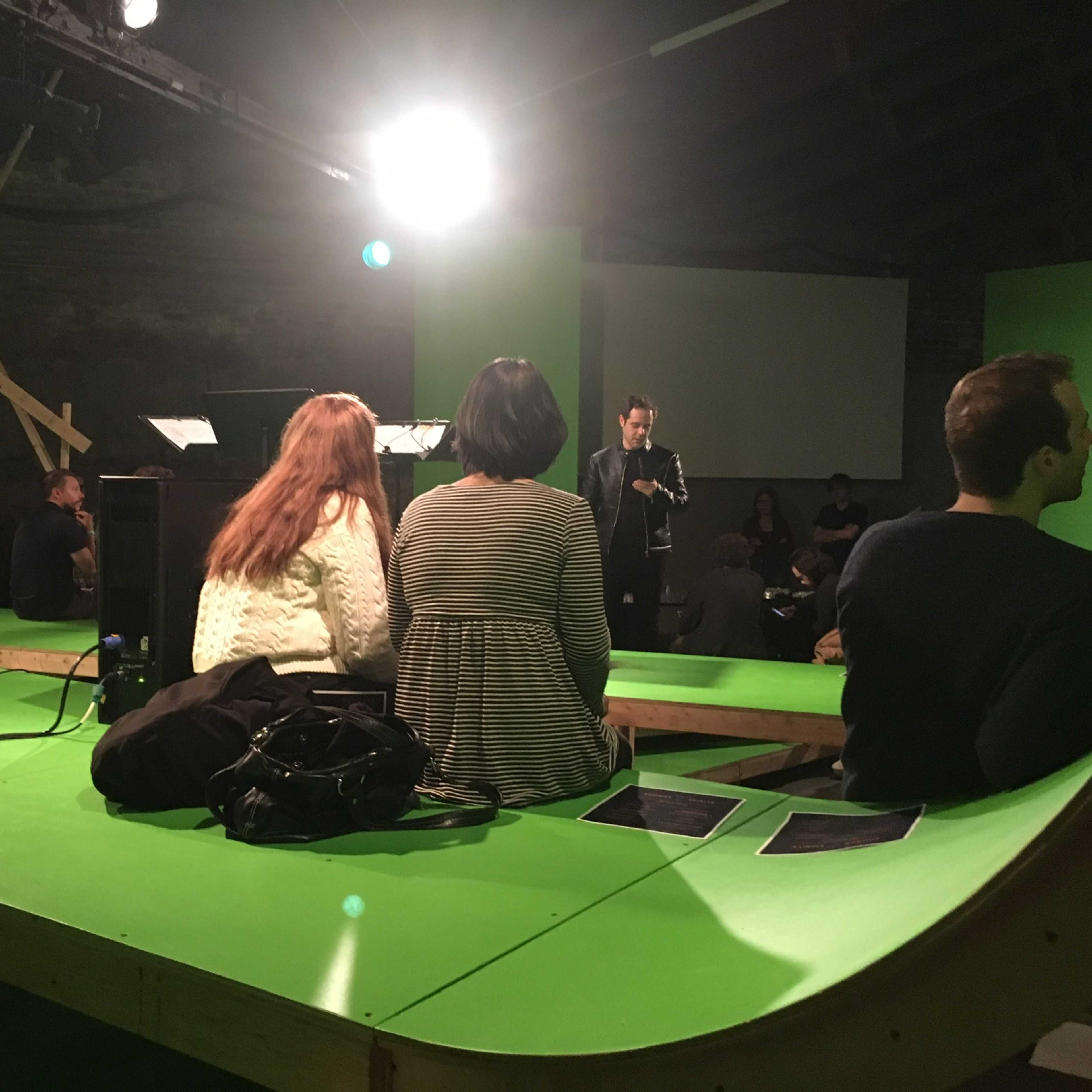 A group of people sits on a green stage with a speaker presenting in front of them.