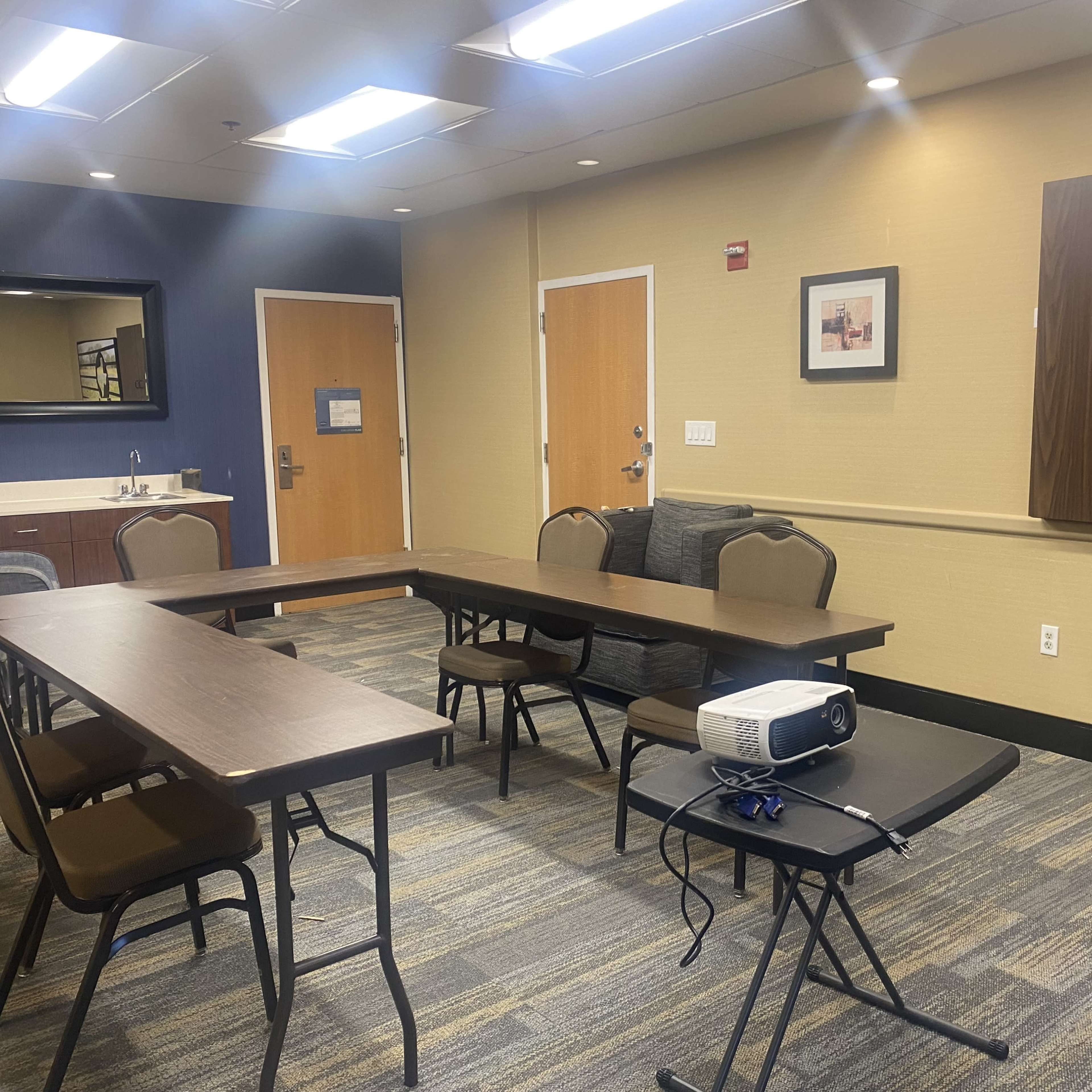A meeting room with a U-shaped arrangement of tables and chairs, a projector on a small table, and a kitchenette area in the background.