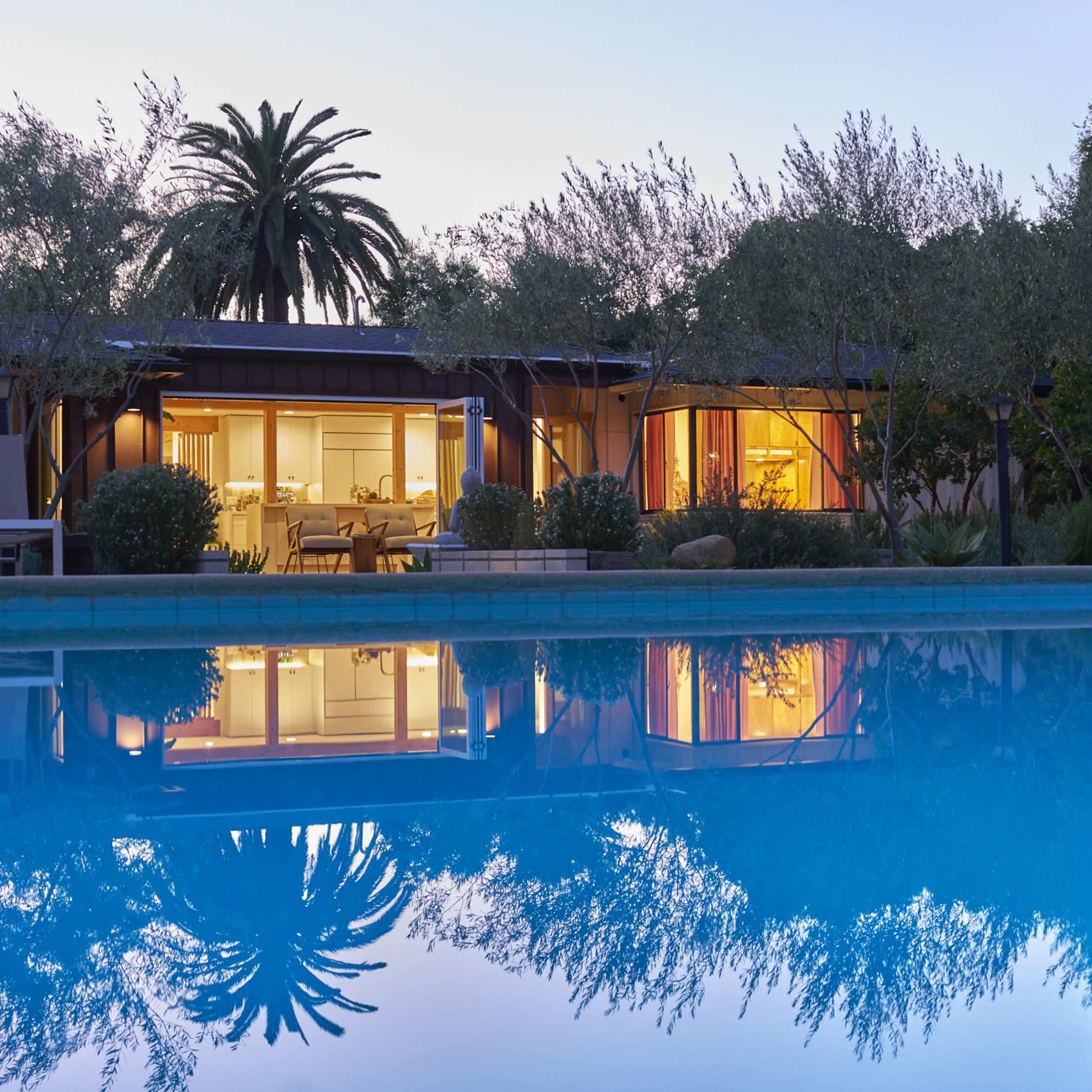 A modern home with large windows overlooks a still pool reflecting the surrounding palm trees and evening sky.