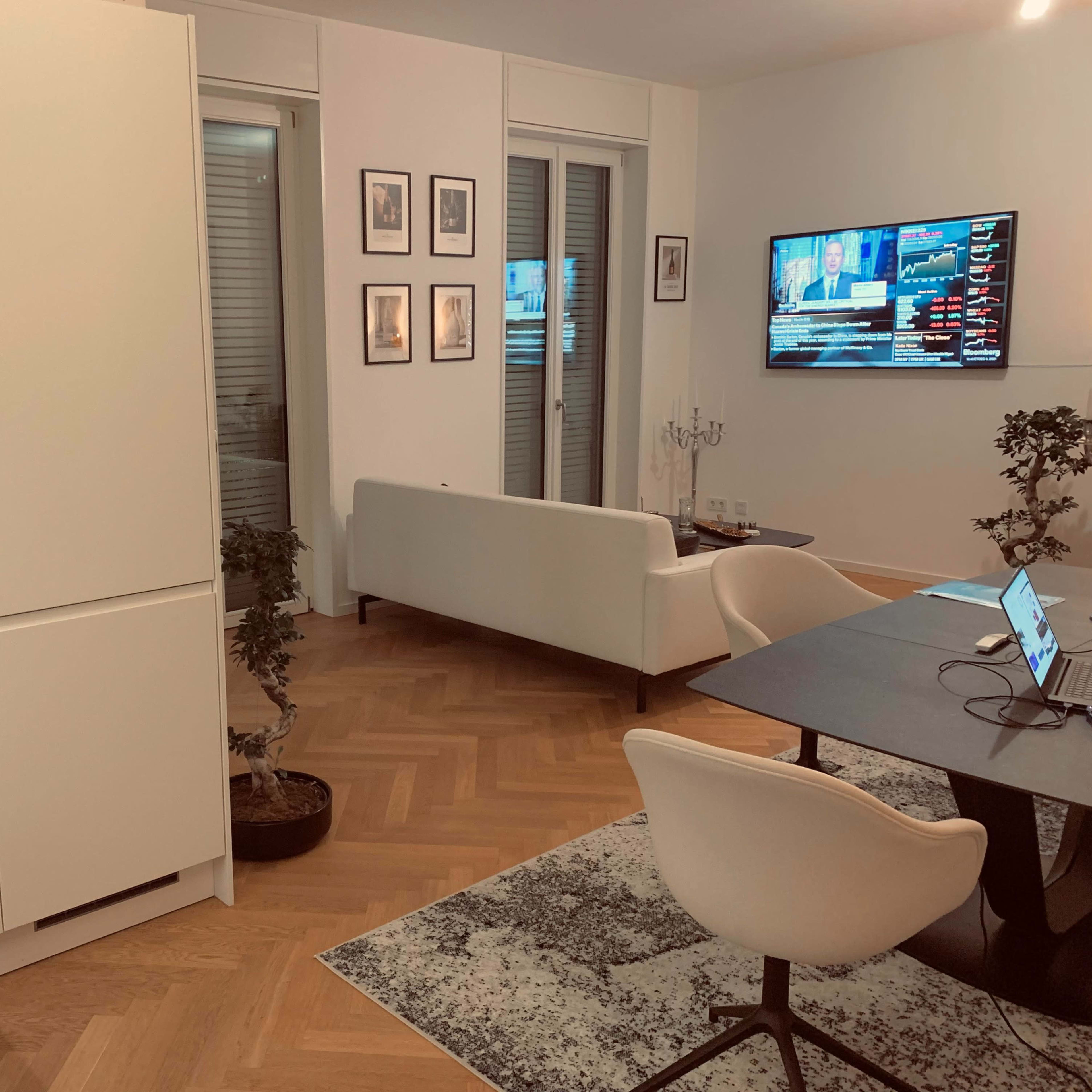 A modern living space features a white sofa, a dining table with chairs, and an oven in a sleek kitchen, with a television displaying news in the background.