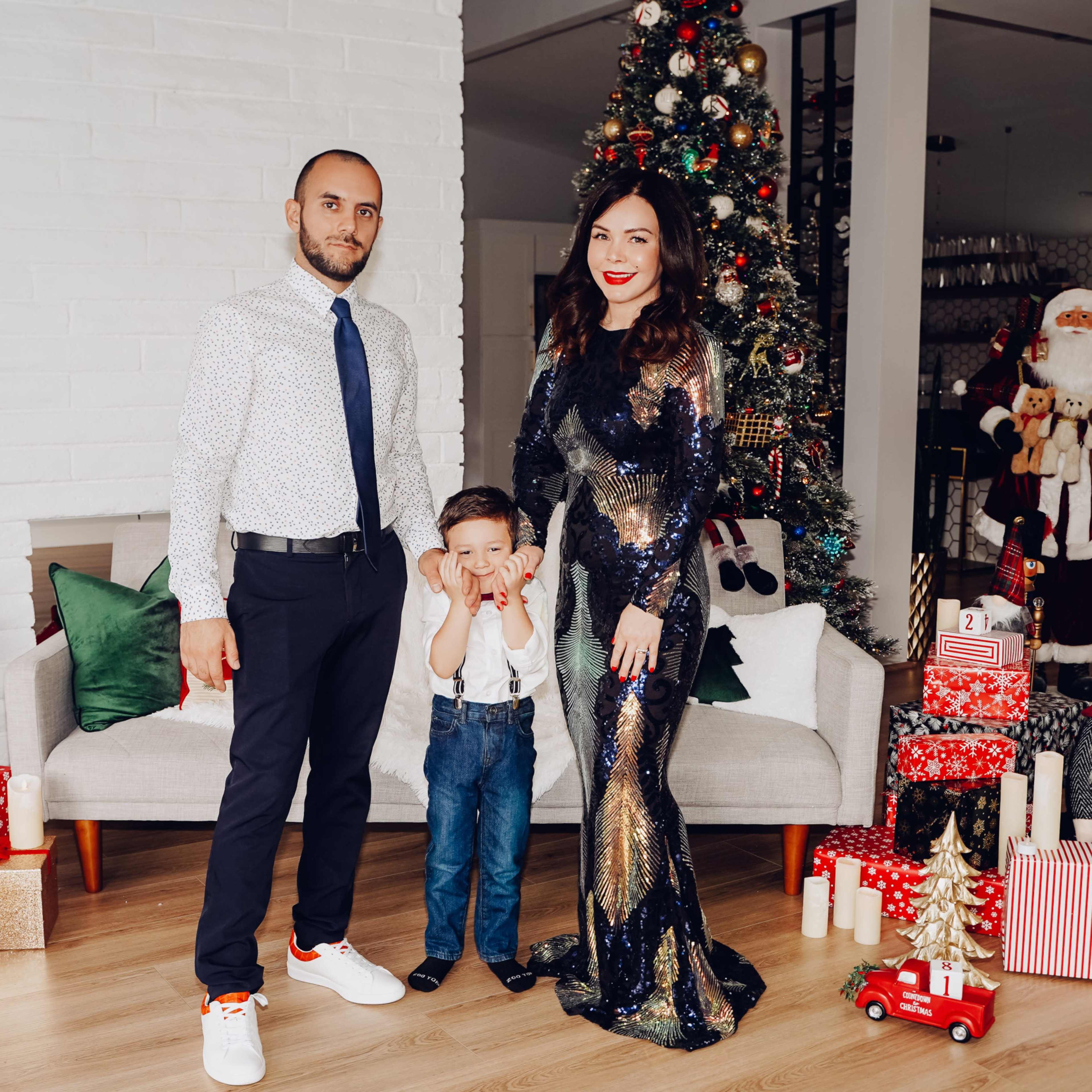 A man, a woman in a sparkling dress, and a young boy in suspenders pose in front of a decorated Christmas tree and festive ornaments.