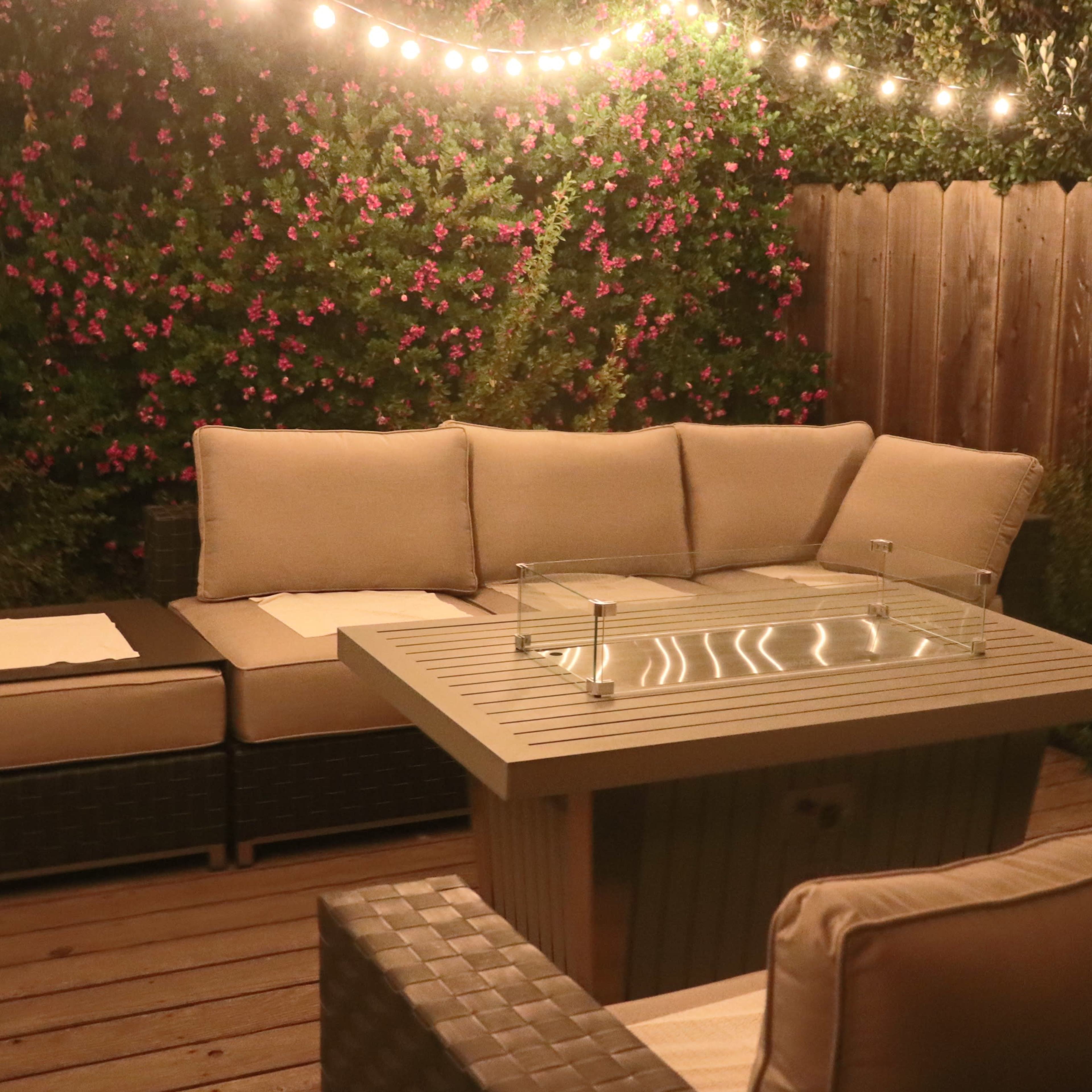 A cozy outdoor seating area features a sectional sofa and a fire pit table, illuminated by string lights against a backdrop of flowering plants.