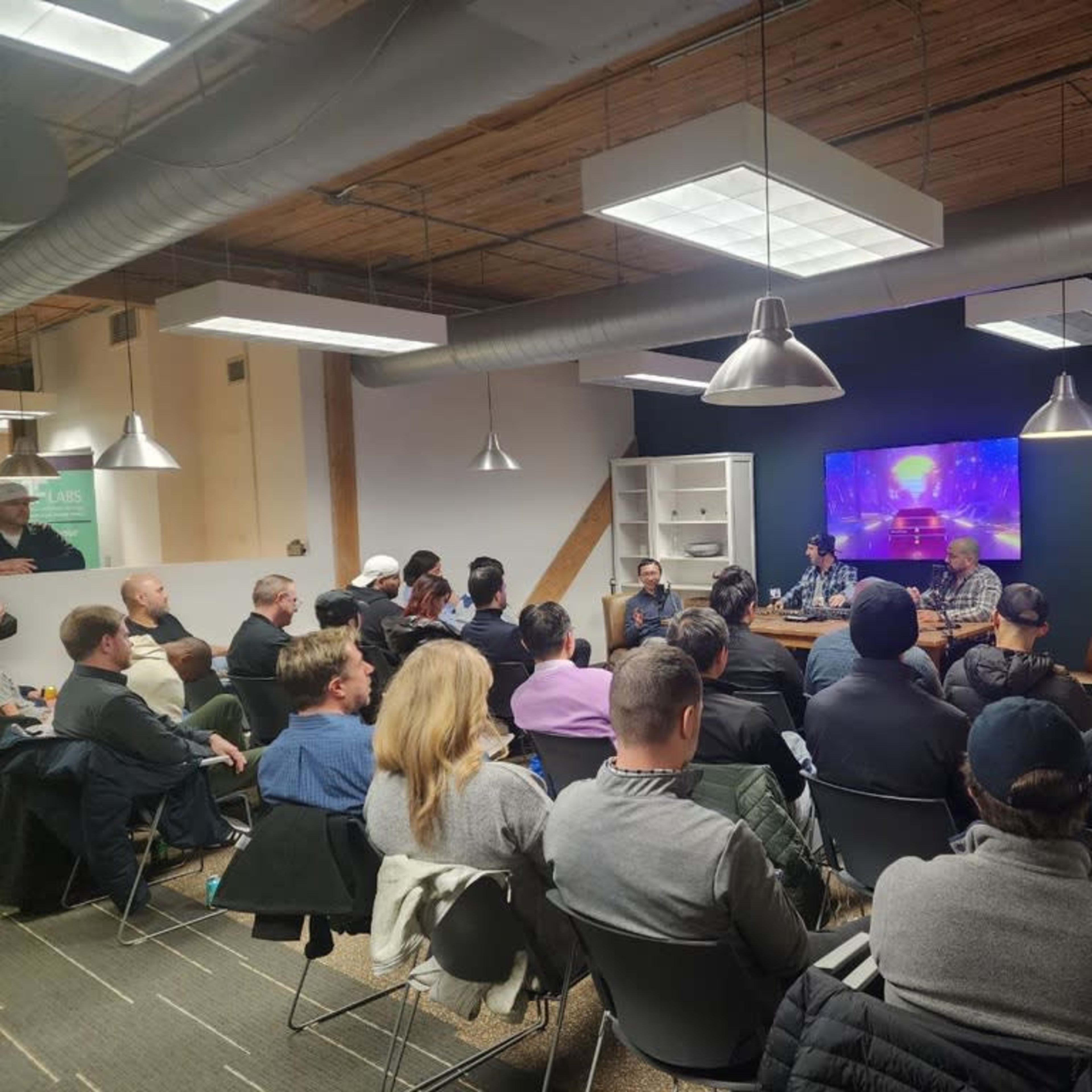 A group of people is seated in a well-lit meeting room, attentively listening to a panel discussion taking place on stage.