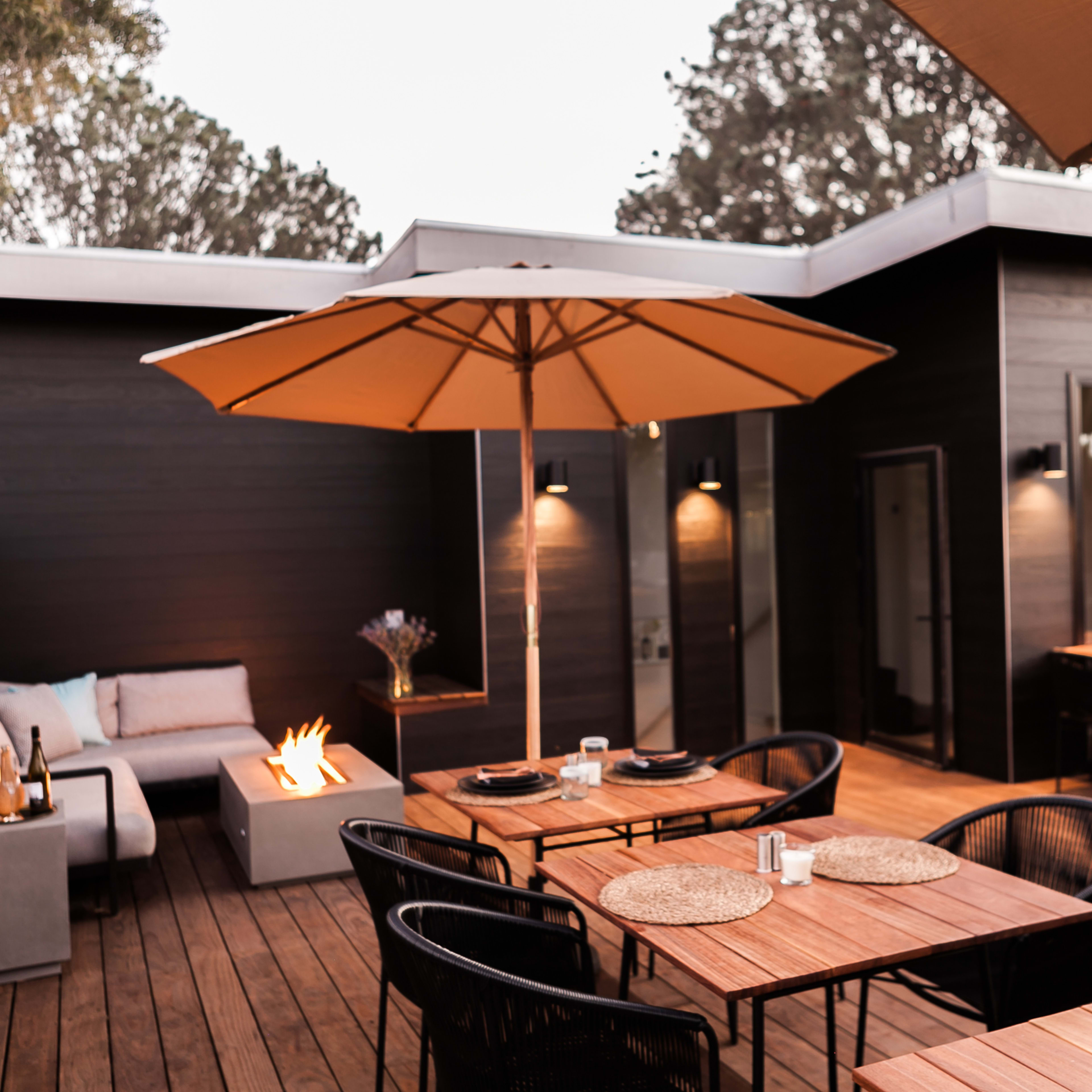 An outdoor patio features wooden decking, dining tables with place settings, a fire pit, and large umbrellas for shade.