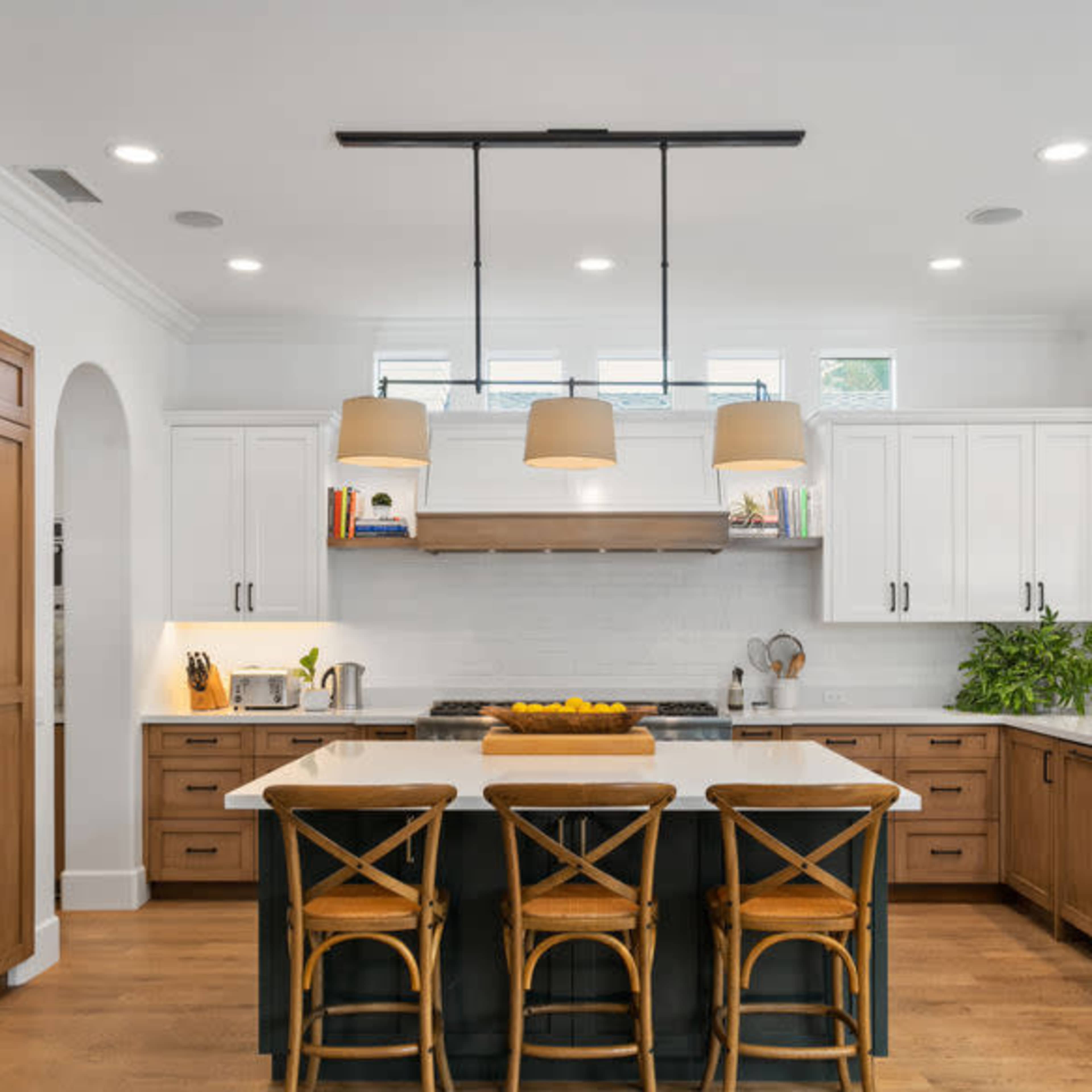 The kitchen features a central island with four wooden stools, surrounded by white cabinets, stainless steel appliances, and pendant lighting.