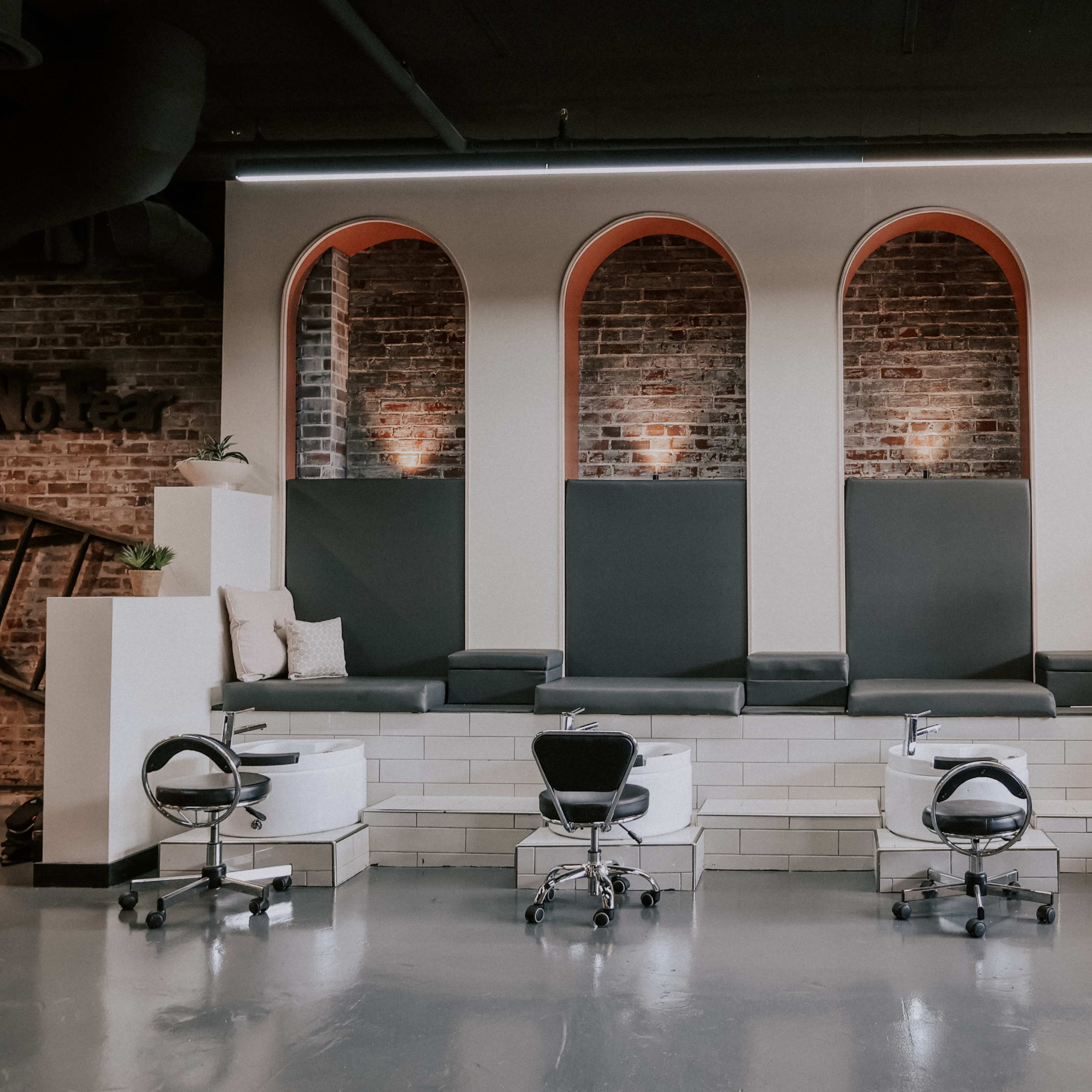 A modern salon interior featuring a series of arched alcoves with seating and pedicure stations, accented by brick walls and minimalist decor.