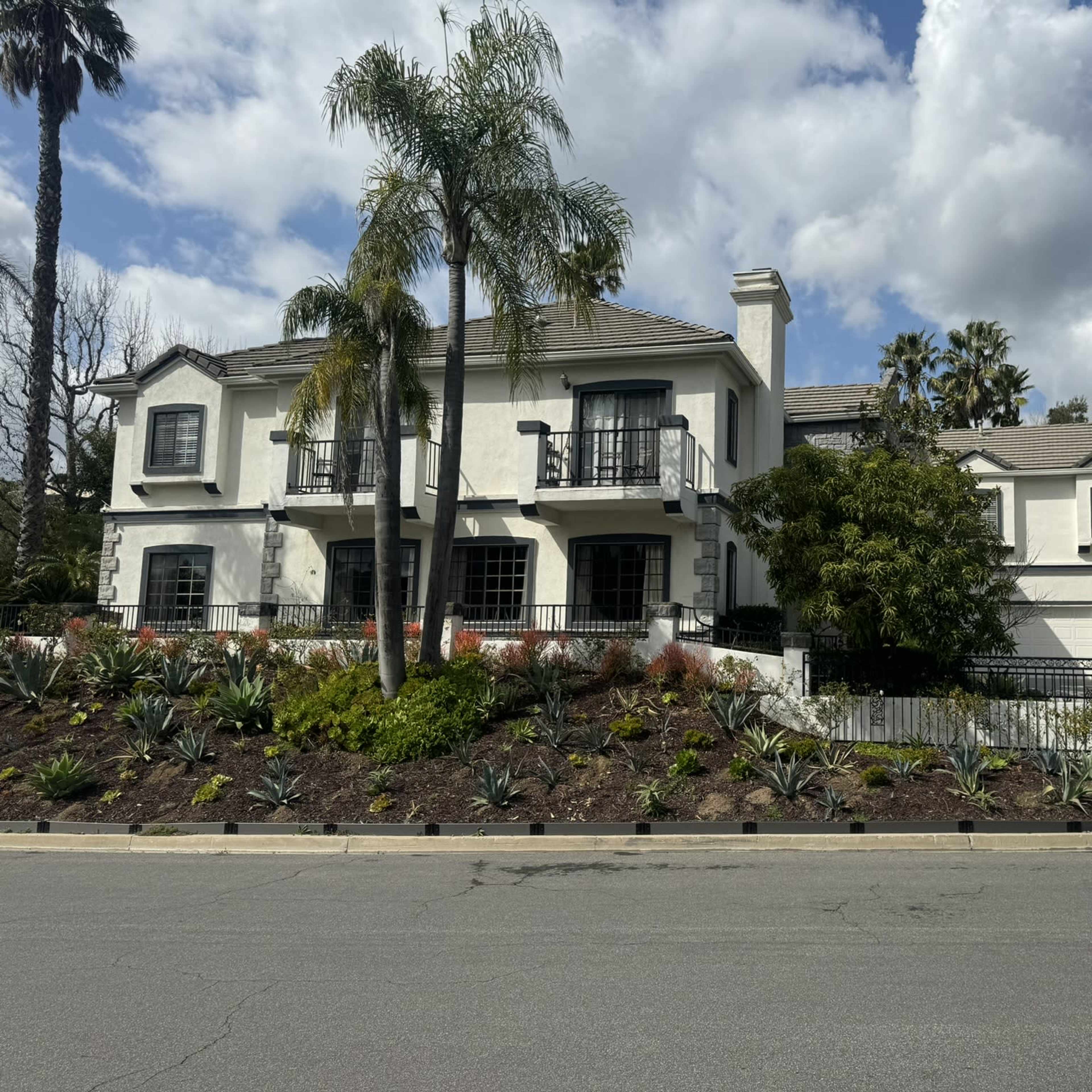 A large, two-story white house with a tiled roof and balcony is set amidst landscaped gardens and palm trees on a suburban street.