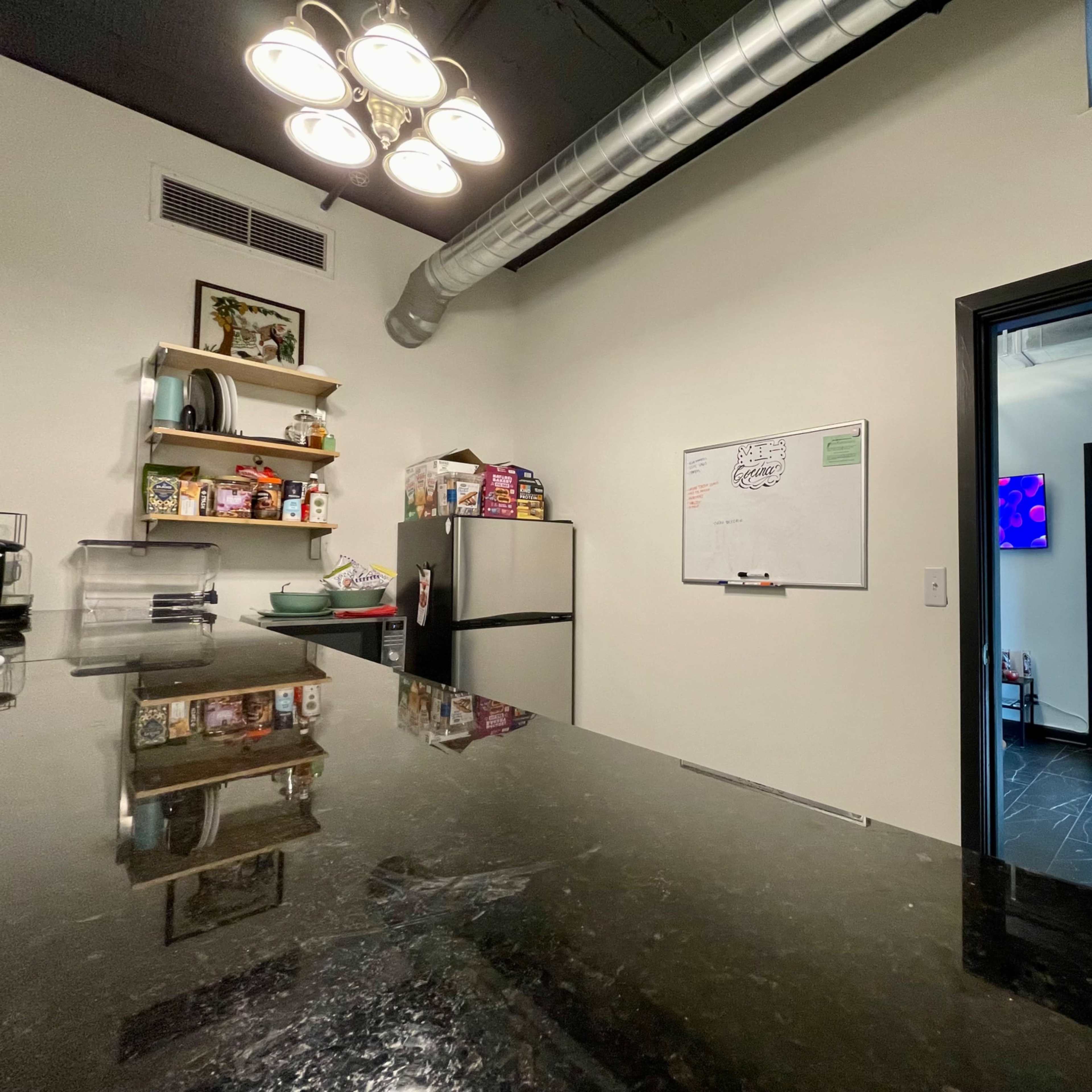 A modern kitchen with a dark countertop, a refrigerator, shelves filled with items, and a doorway leading to another room with a blue light and a stool.