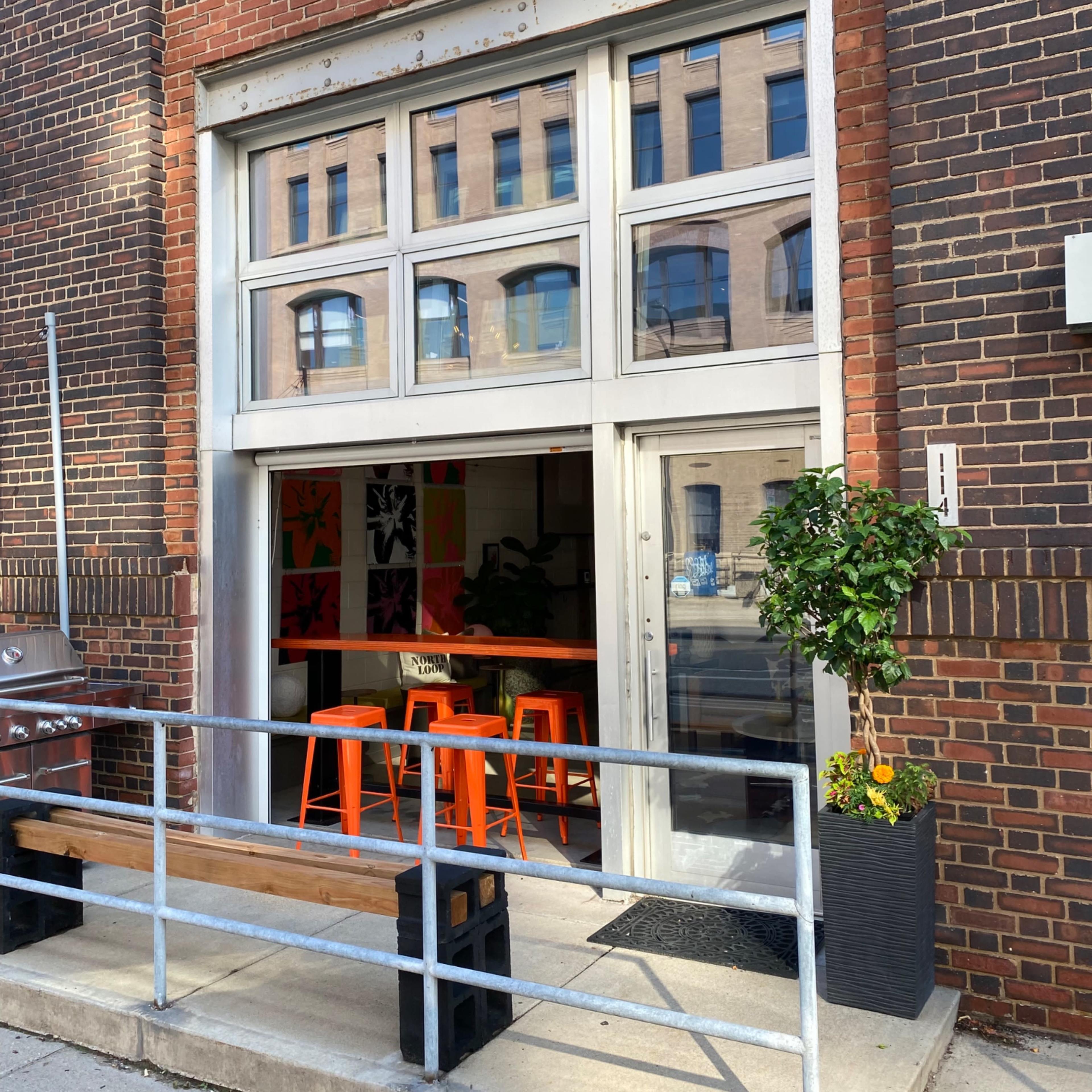 A large storefront with glass doors and a metal railing, featuring orange bar stools visible inside.