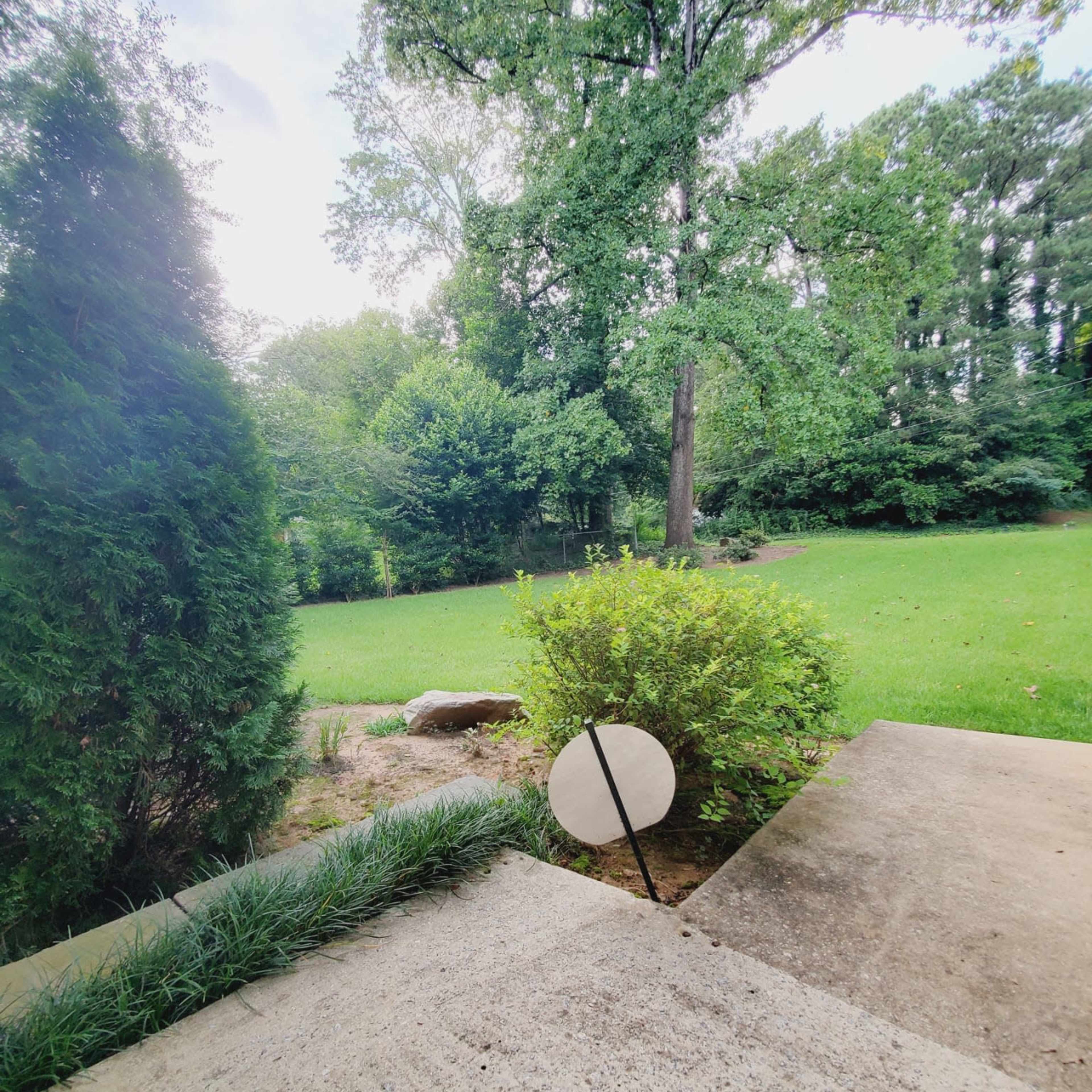 A circular object stands on a concrete surface, with a grassy area and trees visible in the background.