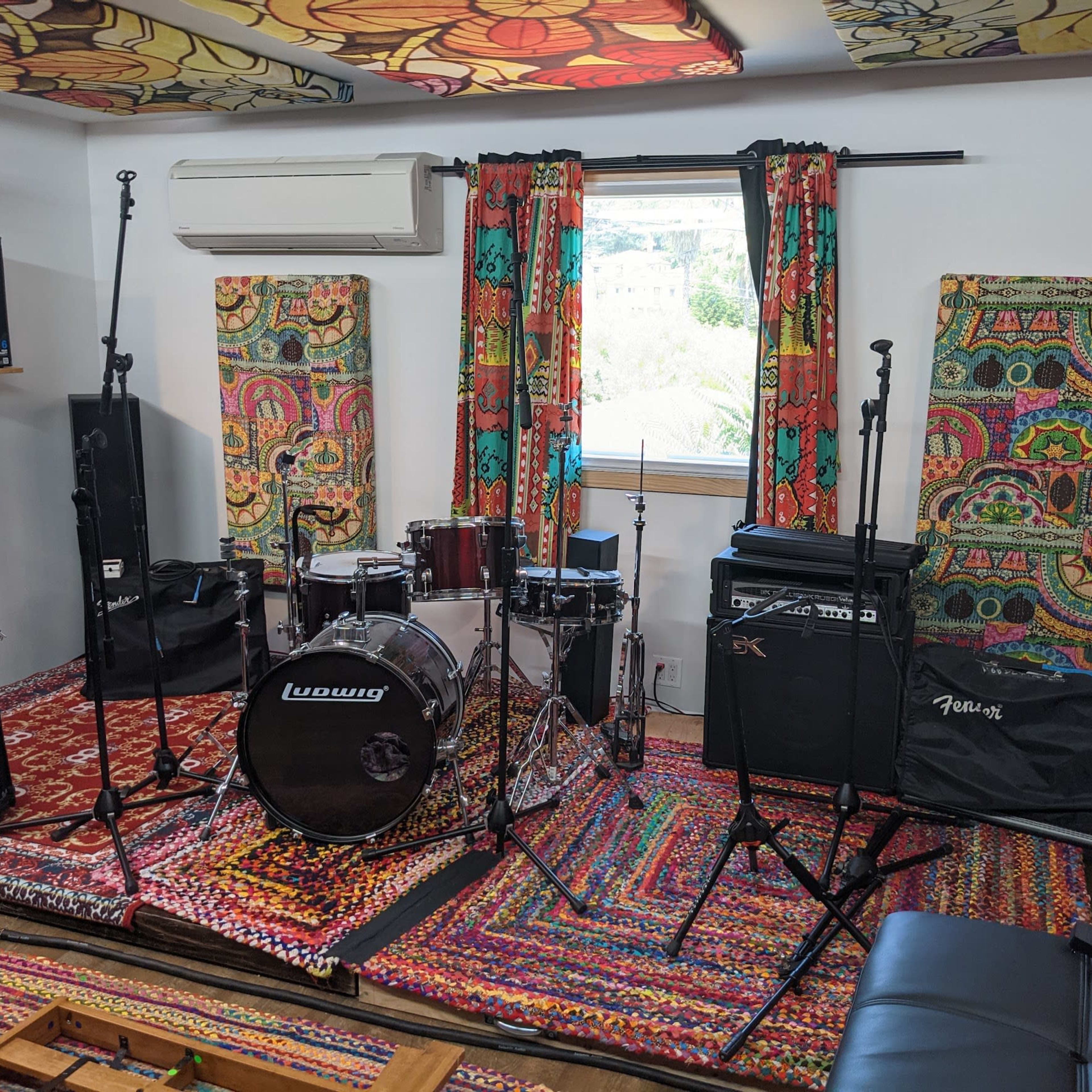 A music rehearsal space with a drum set, amplifiers, microphones, and colorful patterned curtains.