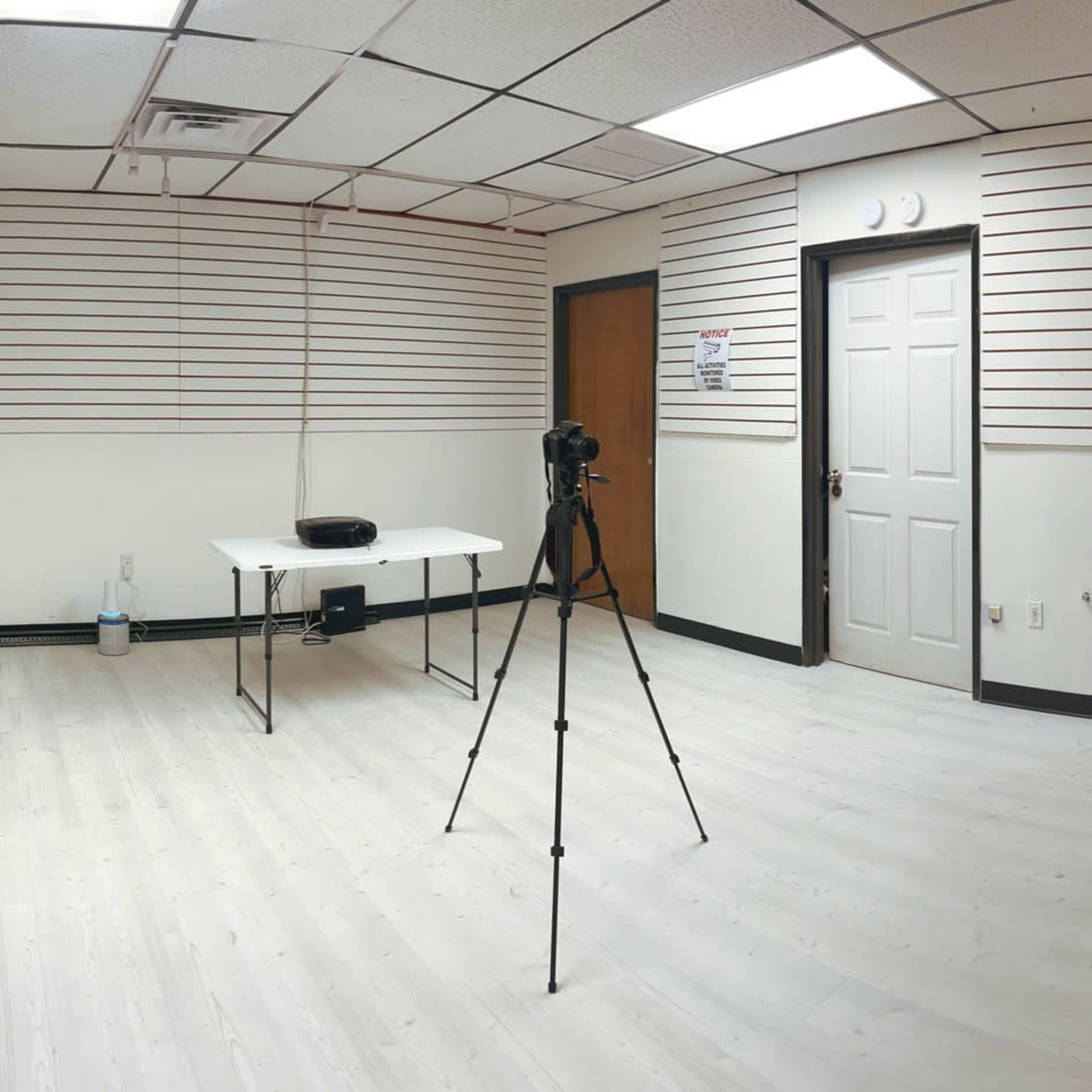 A panoramic view of a sparsely furnished room with a white floor, a table and camera setup, and plain walls featuring horizontal lines.
