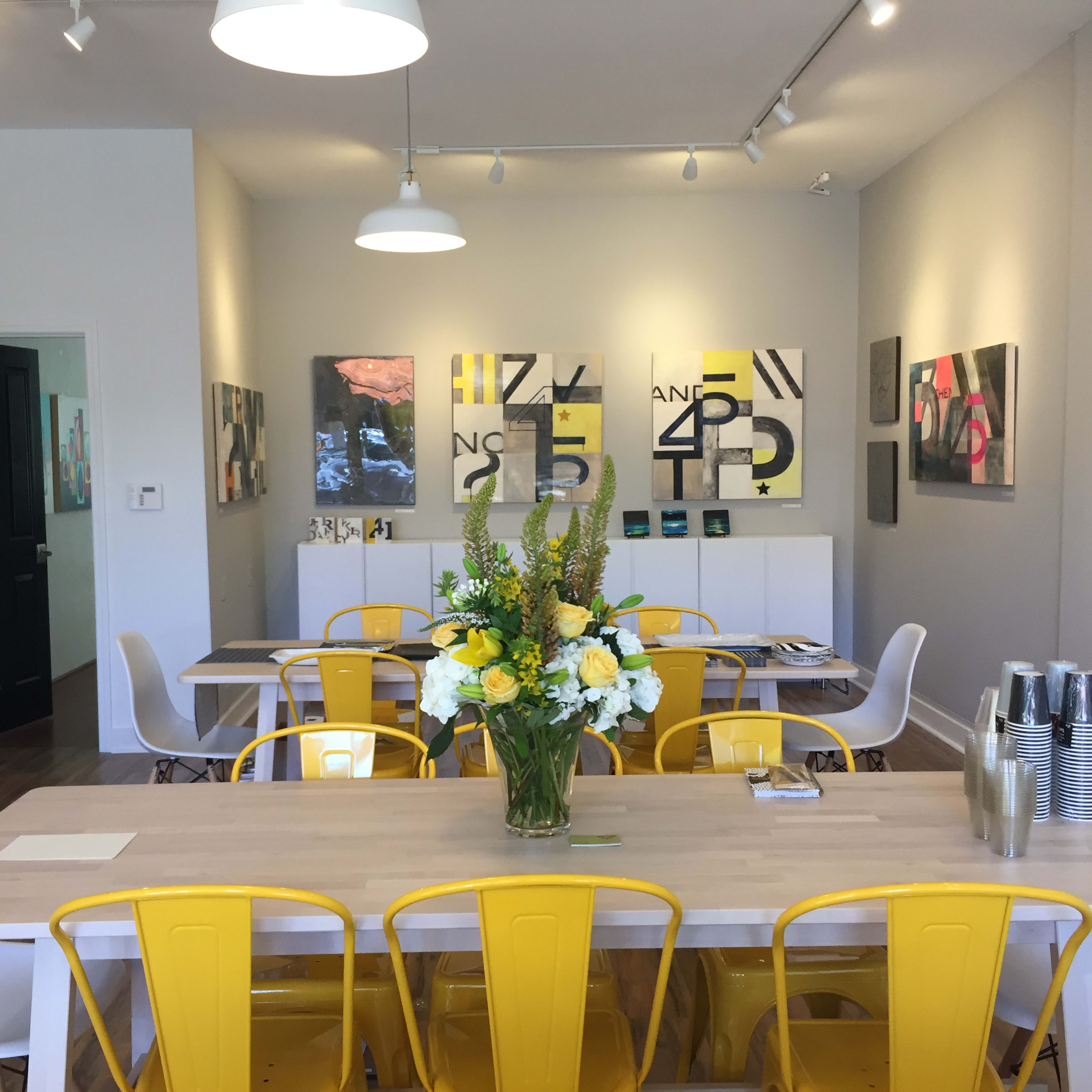 A dining area features a long wooden table surrounded by yellow and white chairs, with artwork on the walls and a floral centerpiece.
