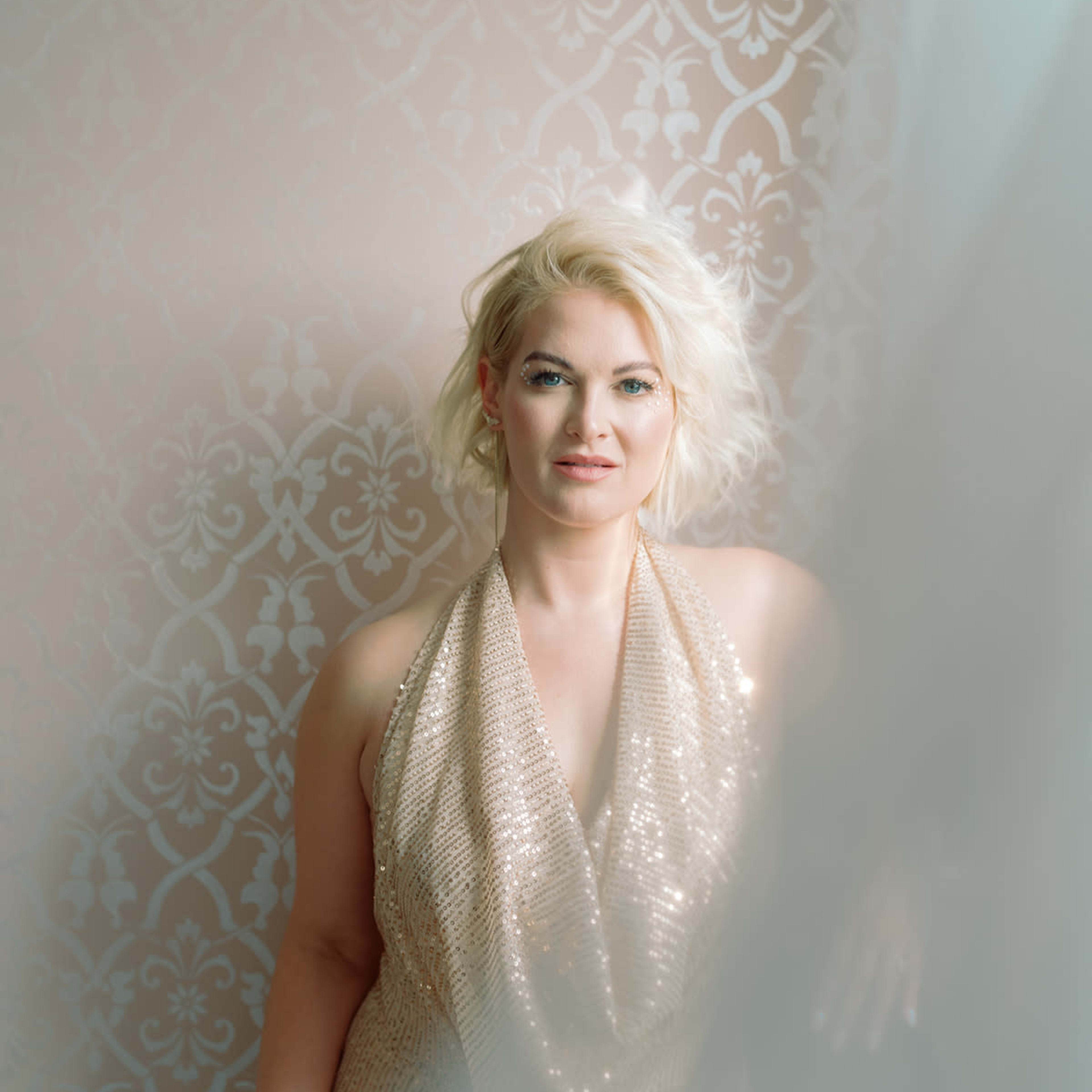 A woman wearing a sequined gold dress poses in front of a patterned wall, partially obscured by a sheer curtain.