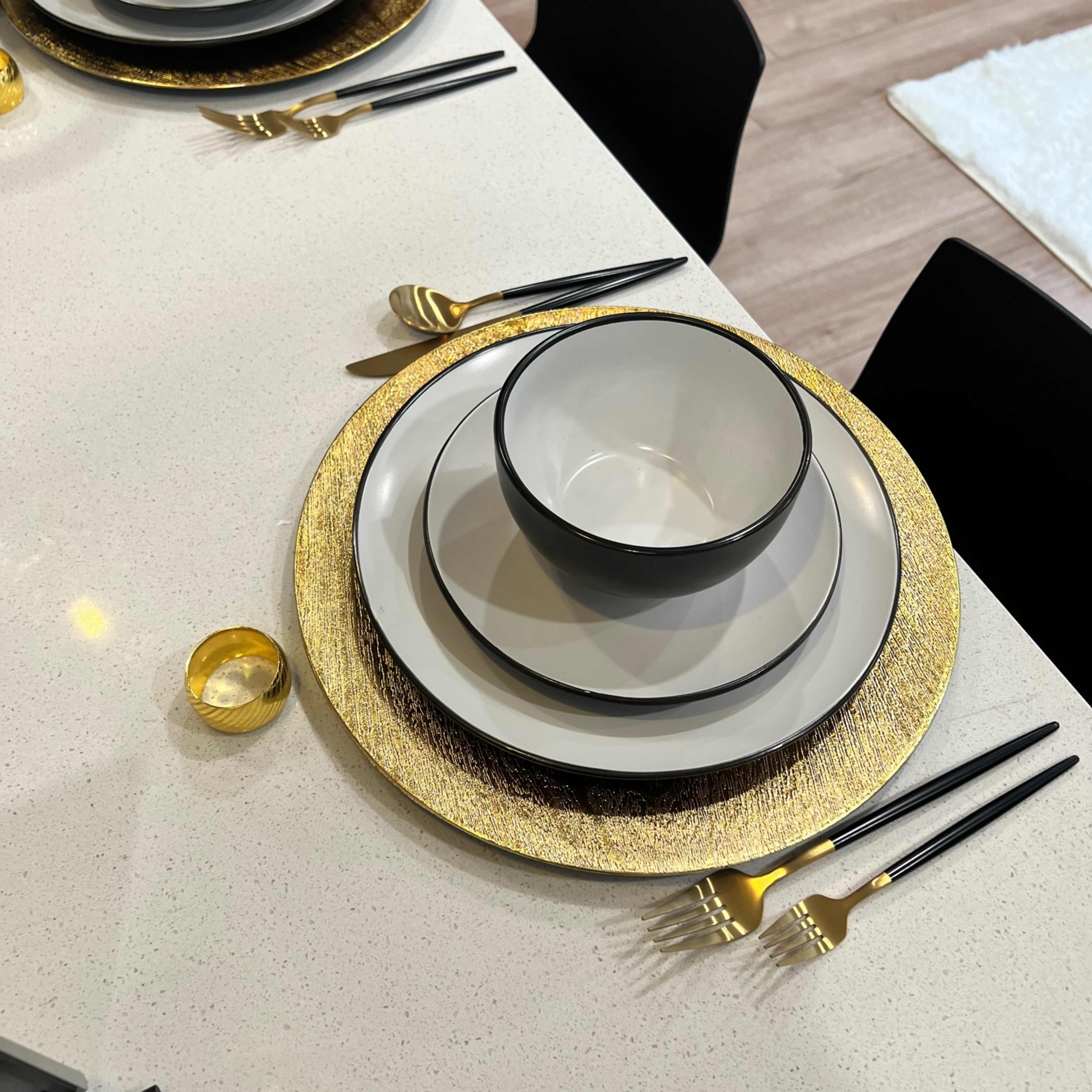 A dining table is set with gold-rimmed plates, black and white bowls, and black utensils on a white surface.