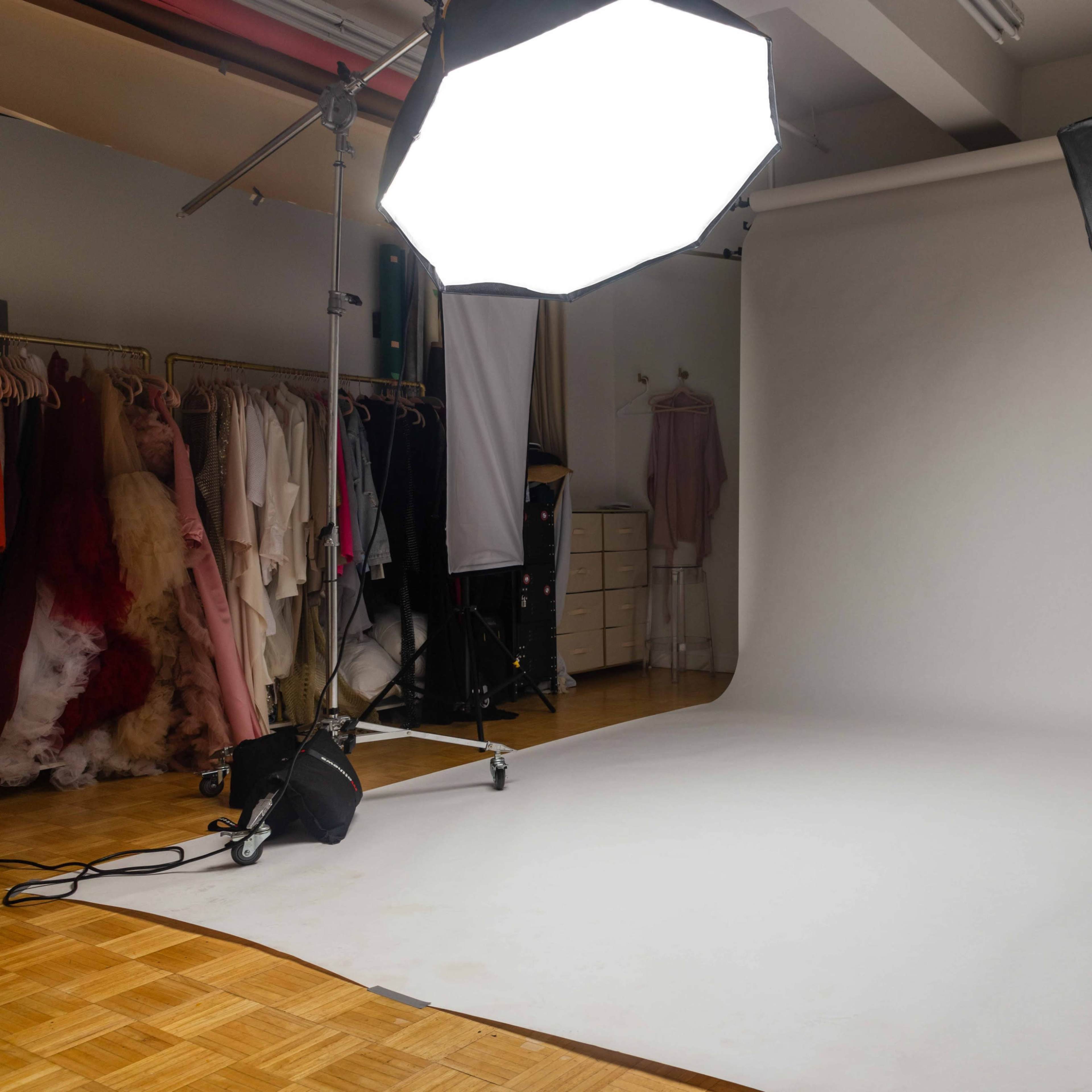 A photography studio setup with a large softbox light, a black umbrella reflector, and a smooth white backdrop, surrounded by hanging garments.