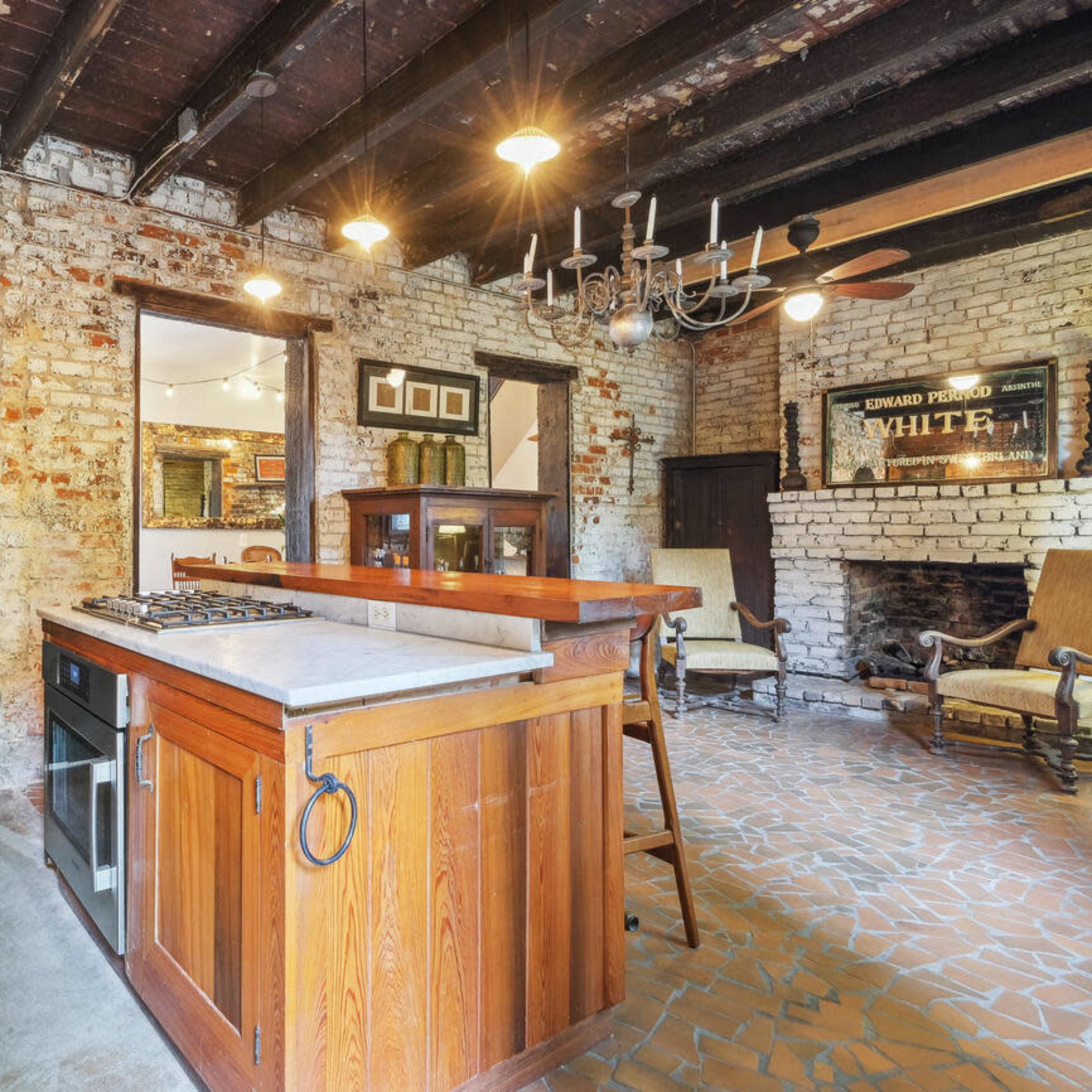 A rustic kitchen featuring exposed brick walls, a central wooden island with a stove, and a chandelier hanging from the ceiling.