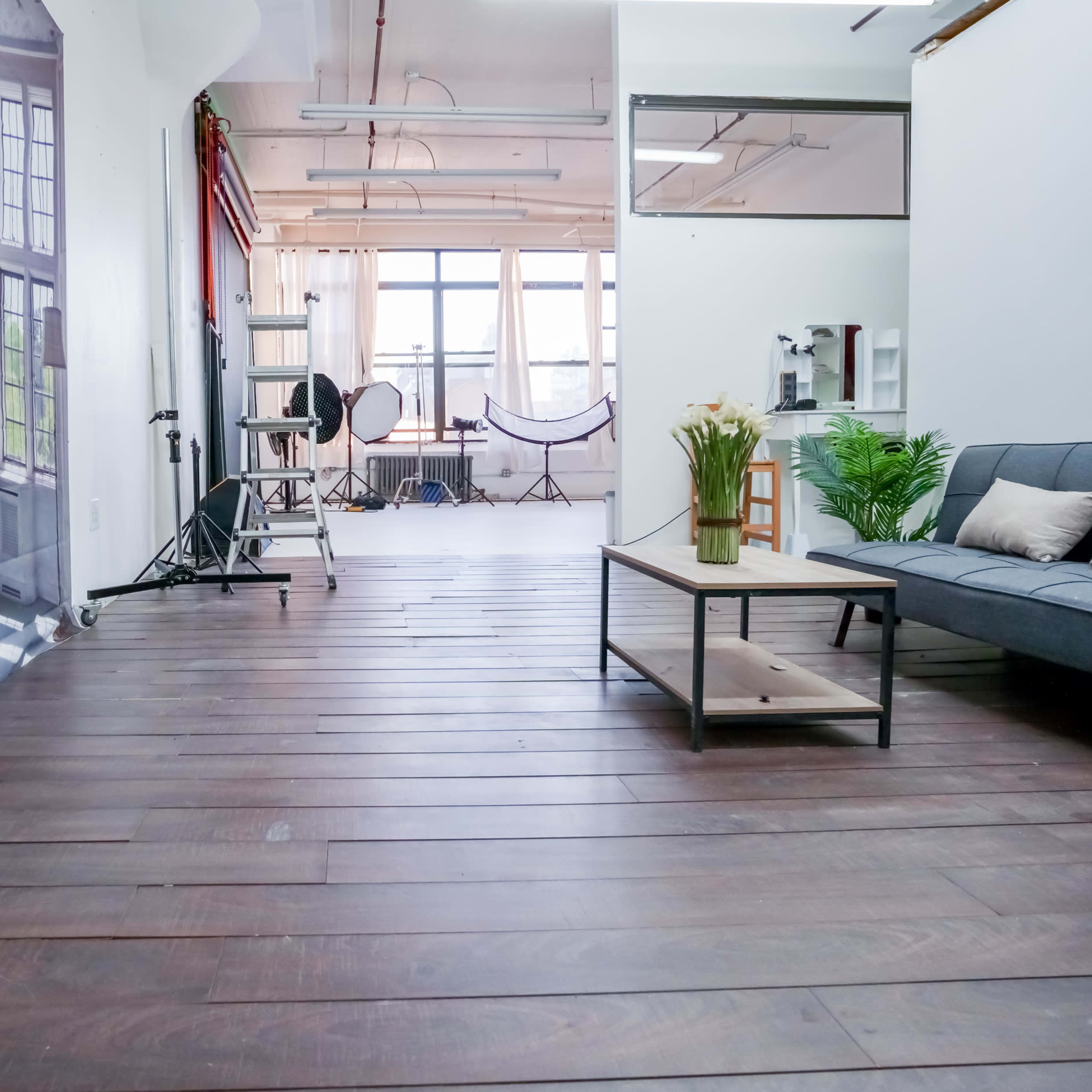 A spacious, well-lit studio featuring wooden flooring, large windows, and photography equipment arranged in the background.