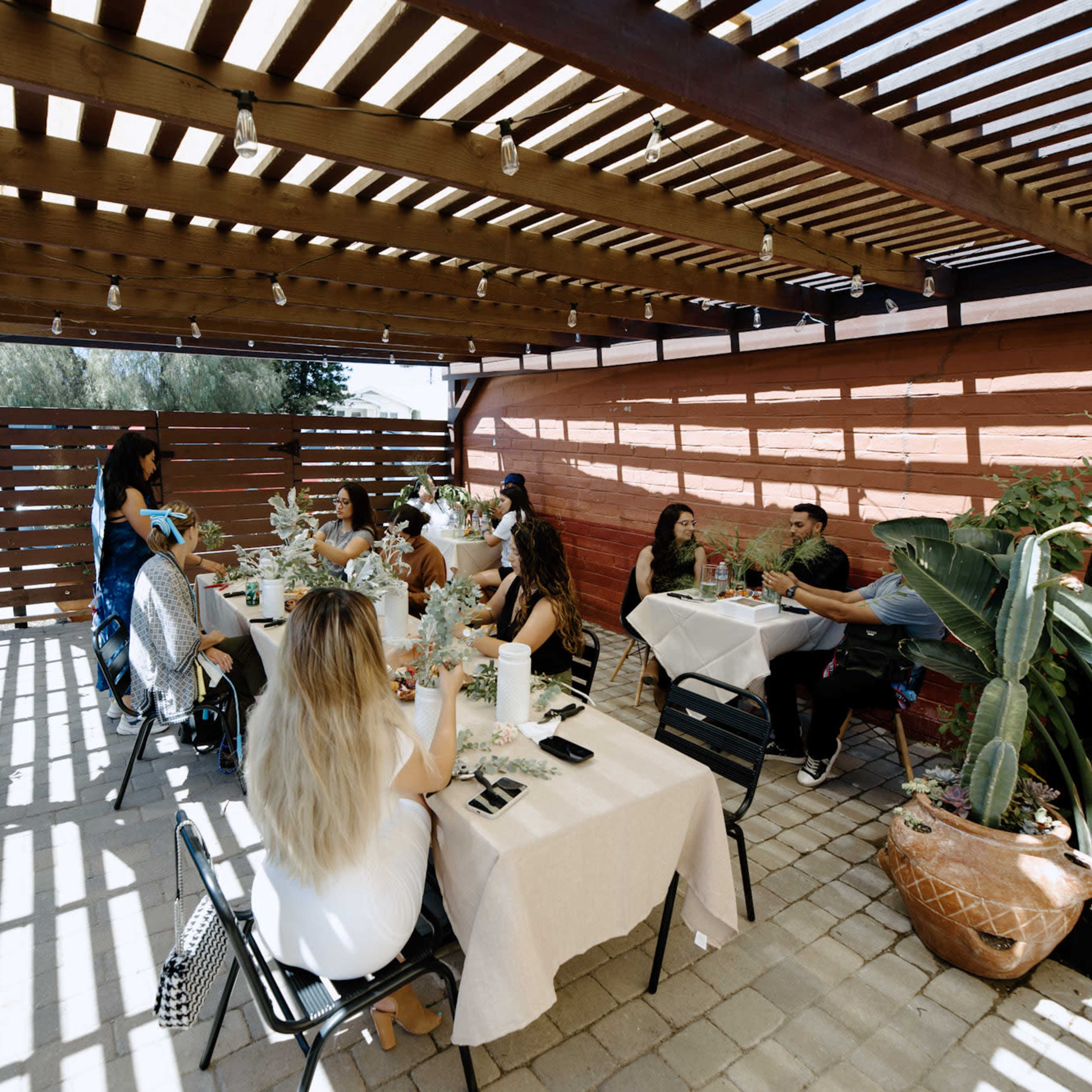 A group of people is seated at tables under a wooden pergola in a shaded outdoor area, surrounded by potted plants.