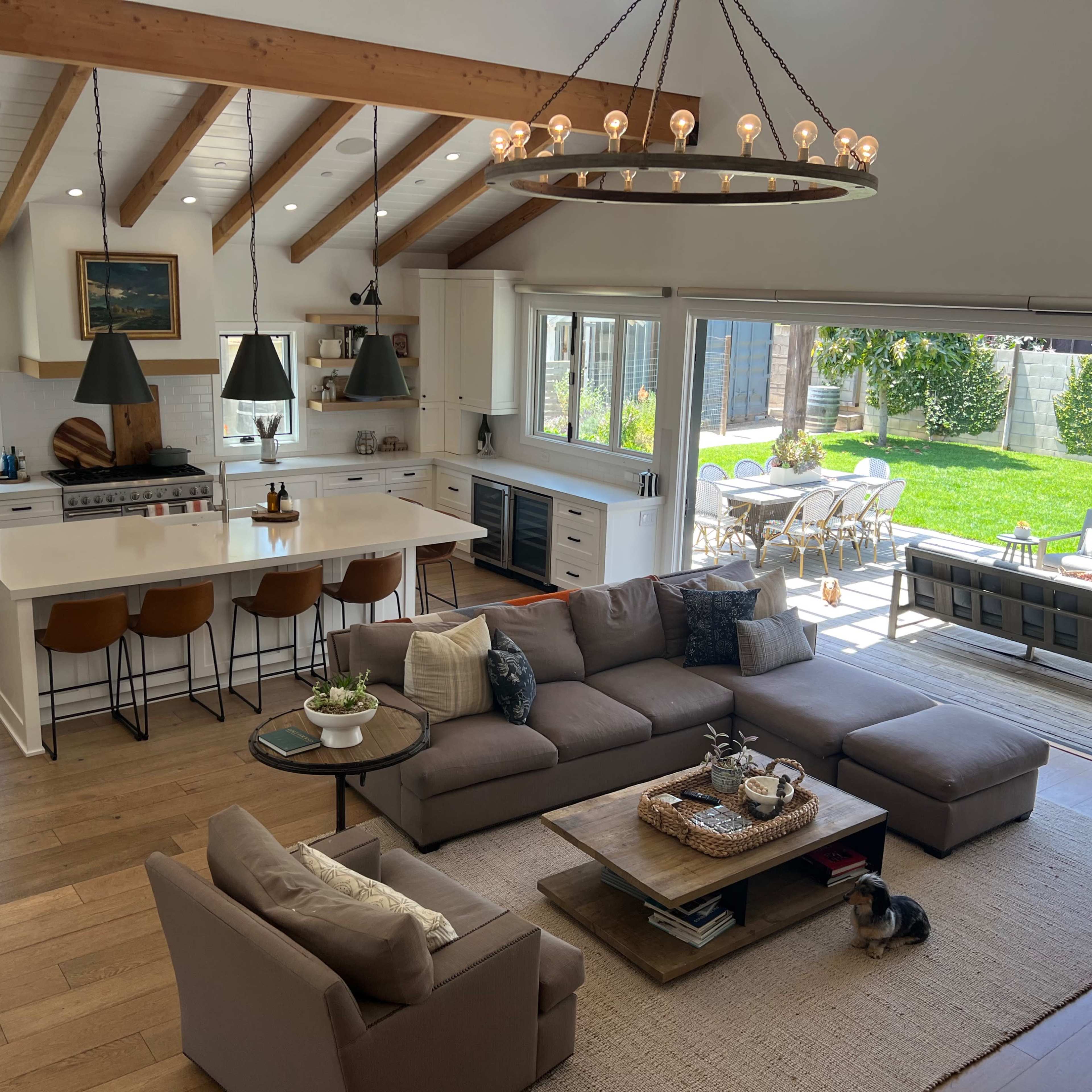 A spacious, modern open-concept living area featuring a kitchen with a large island, a comfortable sectional sofa, and a view of an outdoor dining space with a lush green lawn.