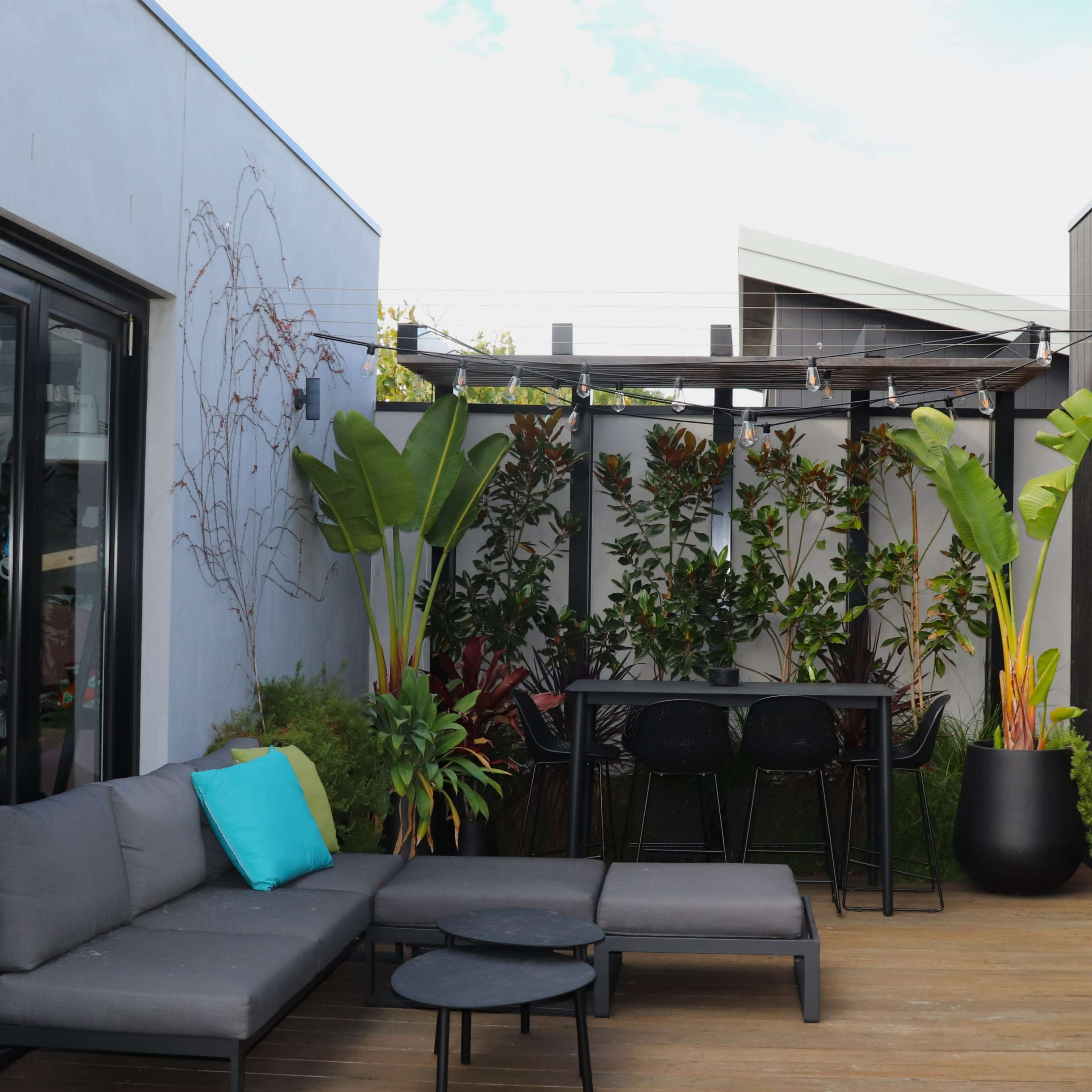 A modern outdoor patio area with a gray sectional sofa, a black dining table surrounded by chairs, and various plants in large pots.