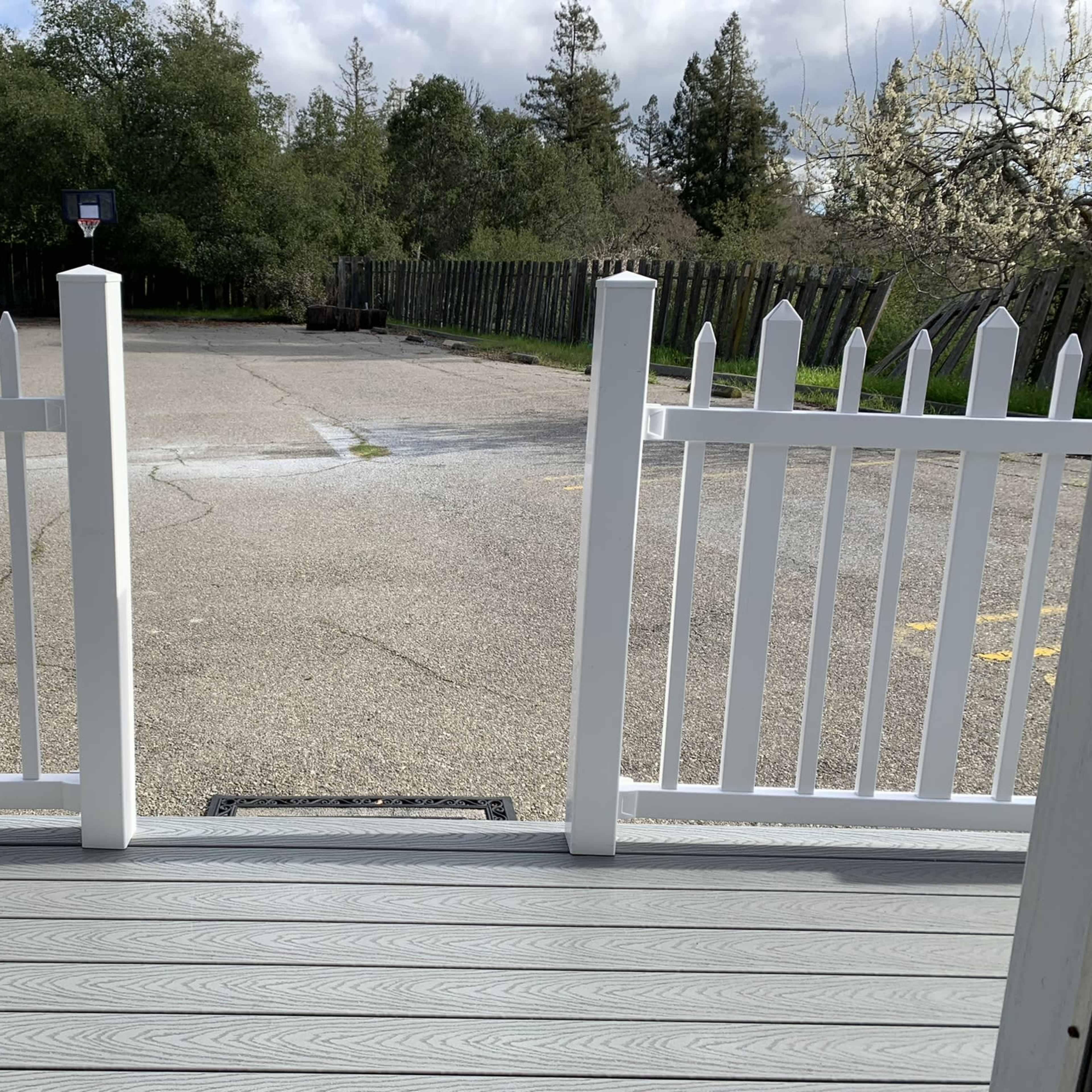 An open white picket fence leading to a gravel parking lot surrounded by trees and a wooden fence in the background.