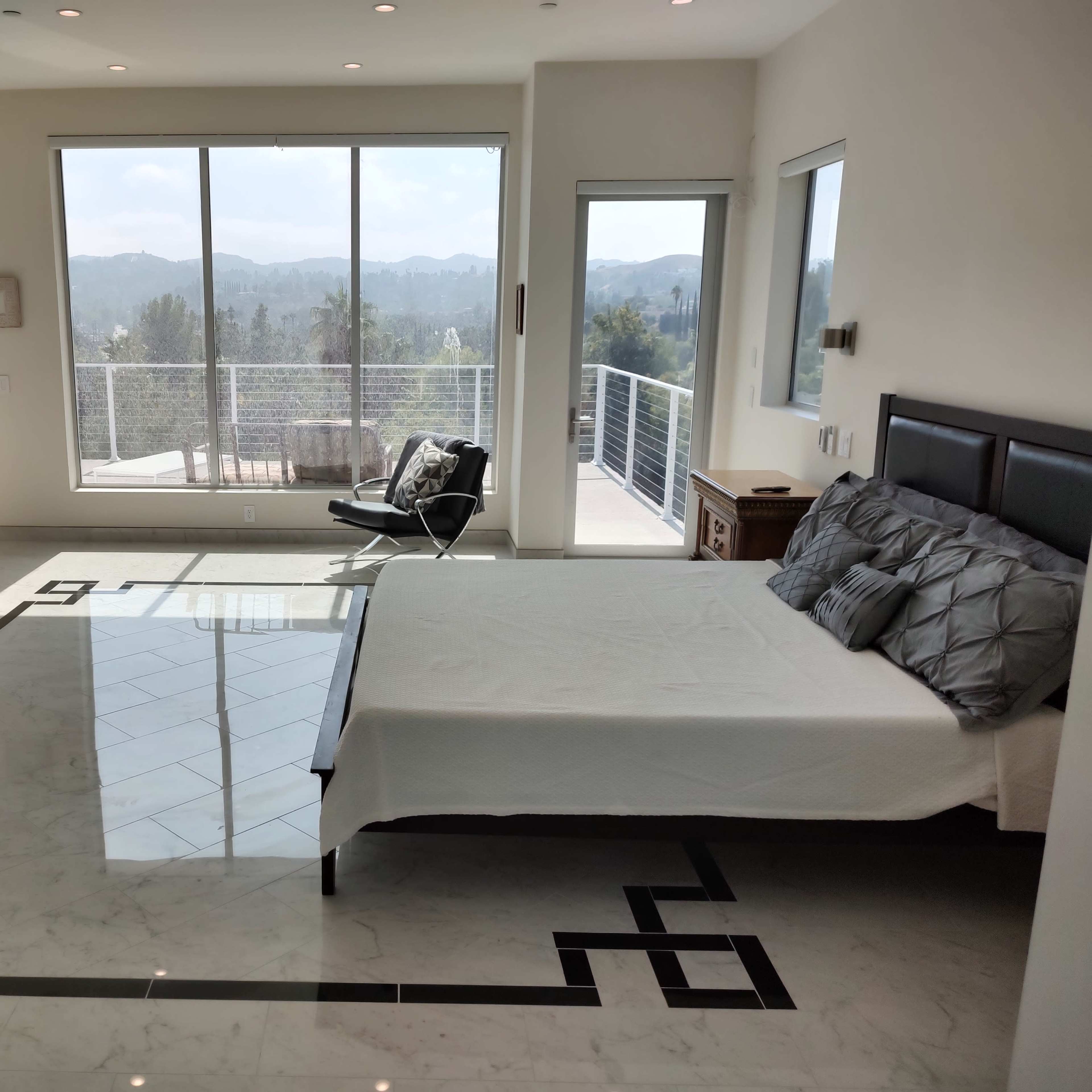 A modern bedroom with a large window that provides a view of mountains, featuring a bed, a chair, and marble flooring with a geometric design.