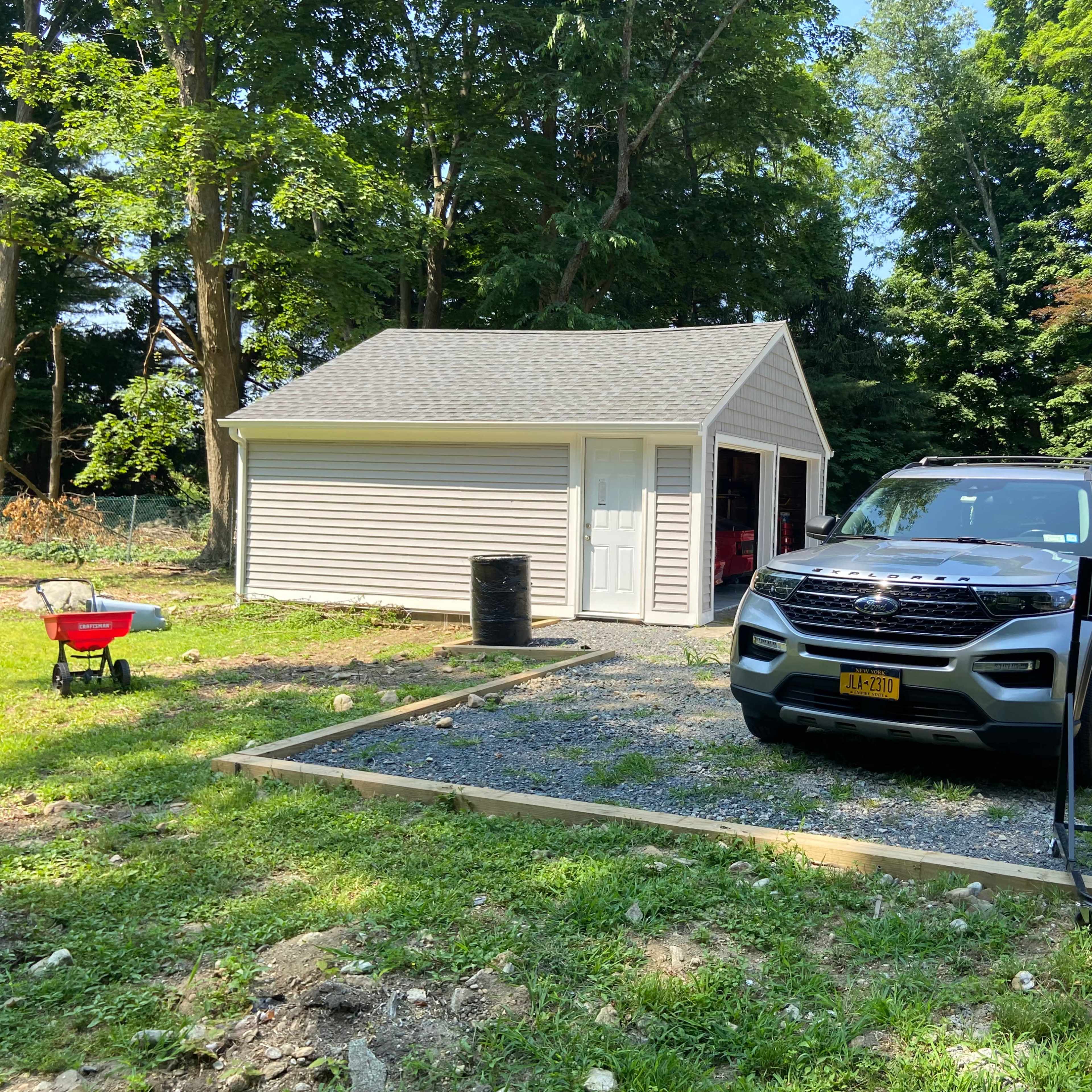 A gray SUV is parked beside a newly constructed garage in a gravel area, with a wheelbarrow and a black barrel nearby.
